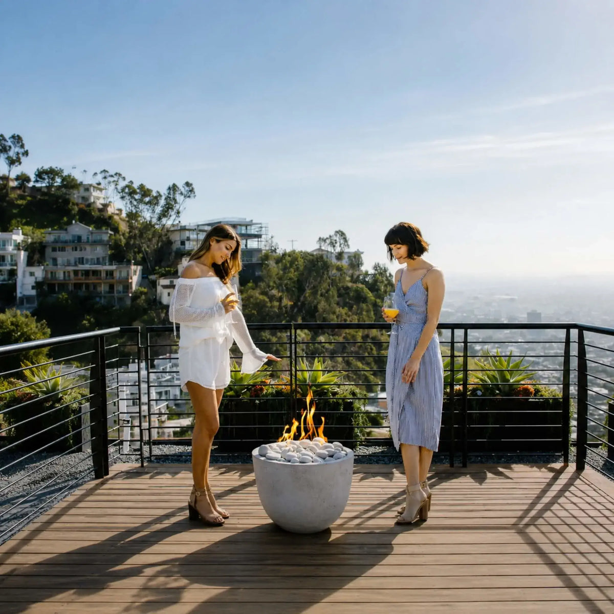 HPC Dekko GFRC Concrete Element Round 20 Fire Column serving as the centerpiece of a contemporary rooftop gathering space. A sleek round concrete fire feature with white decorative stones produces a vibrant flame while guests relax nearby against a panoramic skyline backdrop. The image emphasizes the fire column’s compact footprint, modern styling, and ability to enhance upscale outdoor living environments.