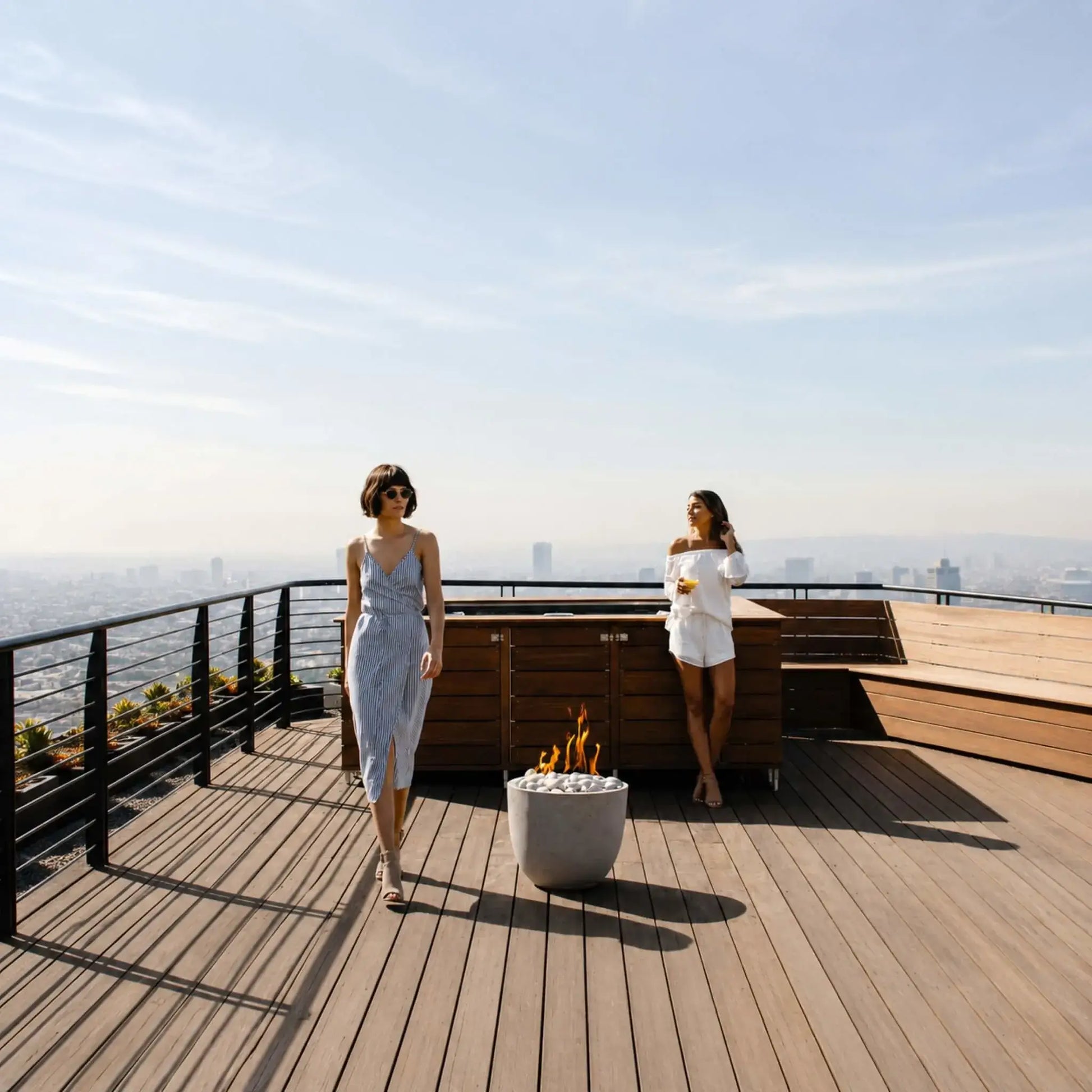 Dekko GFRC Concrete Element Round 20 Fire Column from HPC Fire Hearth Products Controls installed on a spacious wood deck overlooking a city below. The round concrete fire feature is filled with smooth white stones and produces bright flames that contrast with the surrounding metal railing and landscaped planters. This lifestyle scene highlights the fire column’s clean architectural design and outdoor entertainment appeal.