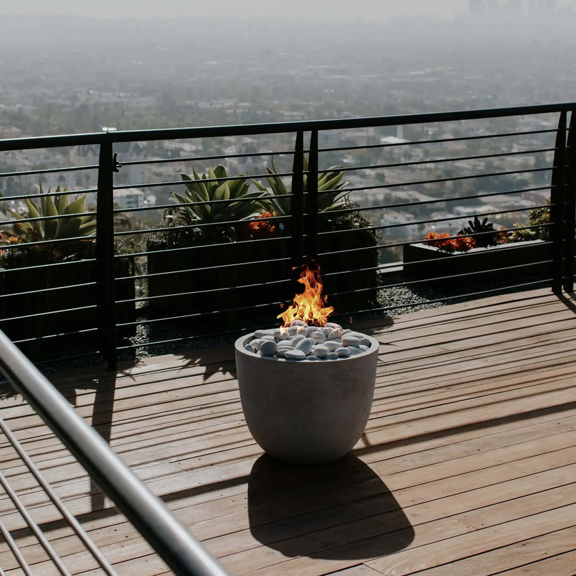 HPC Fire Hearth Products Controls Dekko GFRC Concrete Element Round 20 Fire Column positioned on a modern deck with expansive city views. The compact concrete fire feature burns above polished white stone media, creating a warm focal point against the backdrop of black metal railings, contemporary planters, and a hazy urban skyline. Ideal for luxury rooftop patios and outdoor lounge areas.