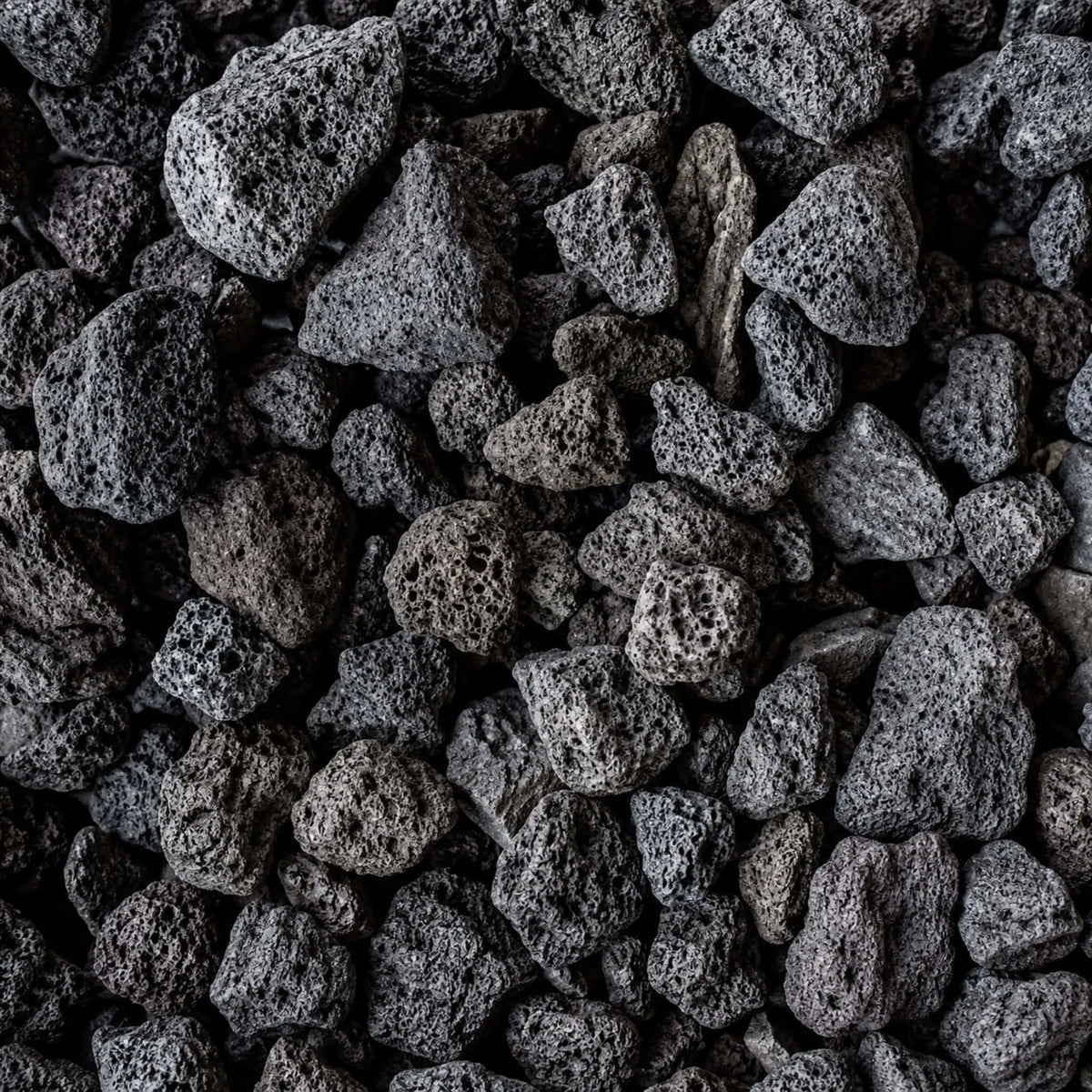 Detailed close-up of HPC Fire Hearth Products Dekko GFRC Concrete Fire Pit Media Lava Rock, showcasing porous volcanic stones with rugged textures and natural black, charcoal, and dark gray coloration. The lightweight lava rock media features irregular shapes and highly textured surfaces designed to enhance flame presentation in outdoor gas fire pits, fire bowls, and custom fire features.
