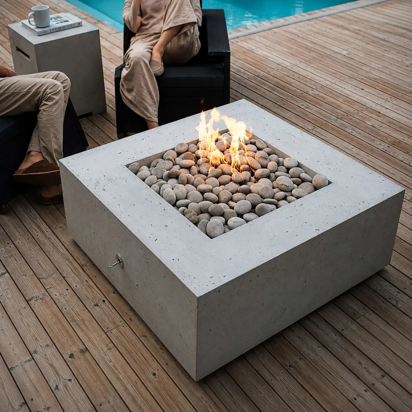 Close-up view of the HPC Fire Hearth Products Controls Dekko GFRC Concrete Propane Tank Cover beside a square concrete fire pit table on a poolside wood deck. The fire pit features decorative river stones and vibrant flames, while the matching propane tank cover doubles as a side table holding a coffee mug and reading material, creating a refined outdoor entertaining space.