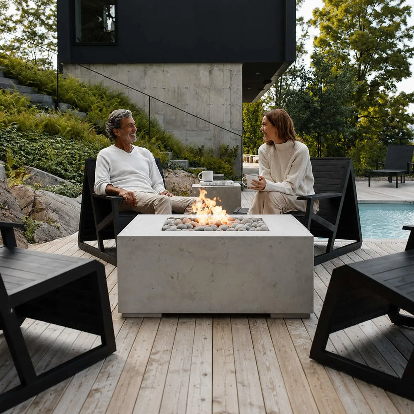 HPC Fire Hearth Products Controls Dekko GFRC Concrete Propane Tank Cover featured alongside a large concrete fire pit on a contemporary poolside deck. Two people enjoy drinks around the fire while surrounded by minimalist outdoor furniture, landscaped hillside greenery, and modern architecture. The matching concrete propane tank cover functions as an elegant accent table while hiding the fuel source.