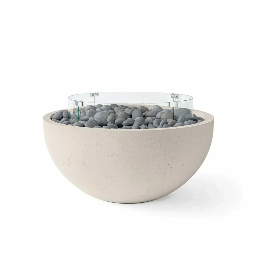 HPC Fire Hearth Products Controls Dekko GFRC Concrete Serenade 32 Fire Pit in Clay finish featuring smooth slate stones and a clear tempered glass wind guard. The rounded GFRC concrete fire bowl blends soft neutral tones with decorative gray stone media, creating an elegant and contemporary outdoor fire feature suited for patios, courtyards, and modern backyard spaces.