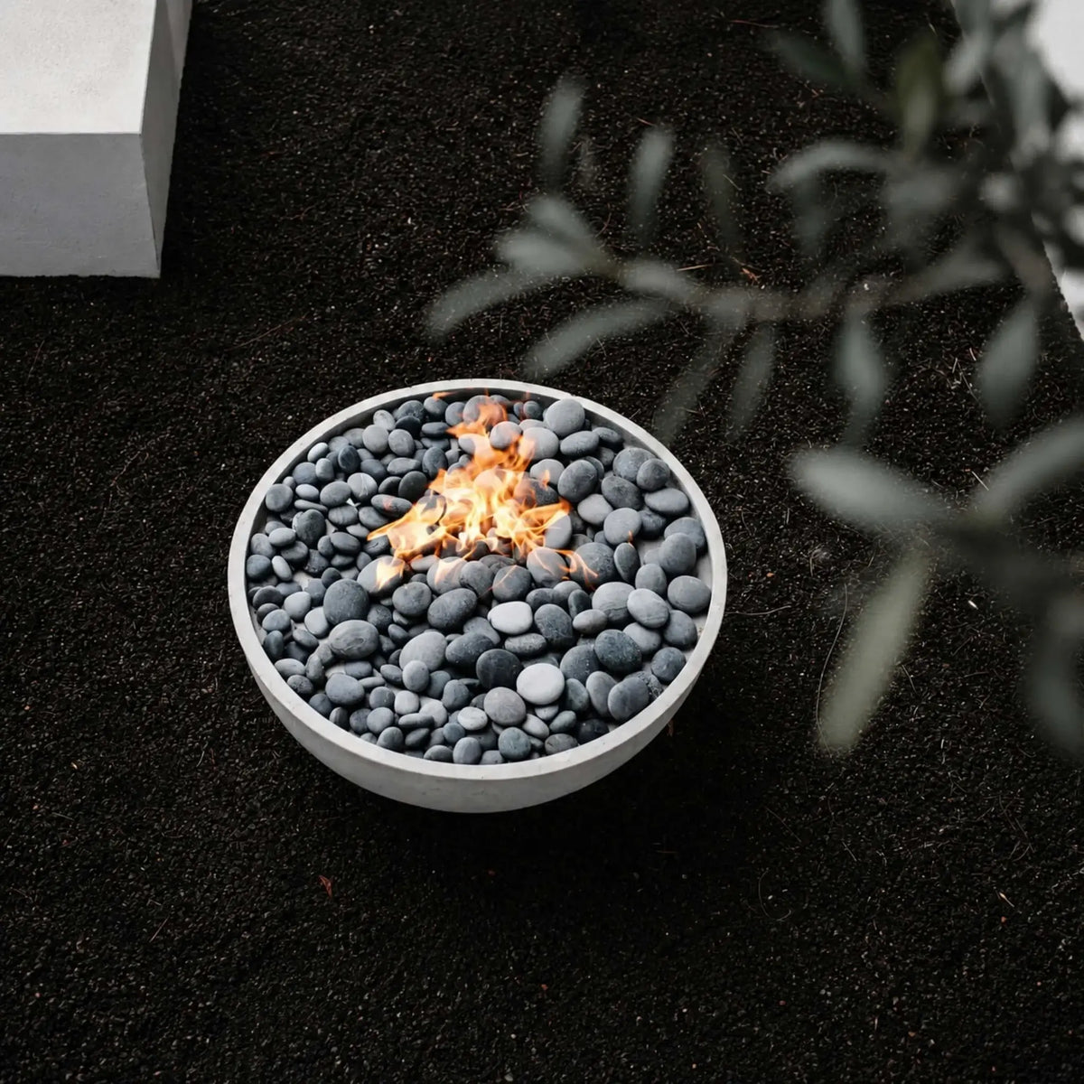 HPC Fire Hearth Products Controls Dekko GFRC Concrete Serenade 32 Fire Pit displayed in a modern landscape setting with smooth gray fire stones and a natural gas flame burning at the center. The round concrete fire bowl sits on dark decorative gravel, viewed from above, while soft-focus greenery frames the foreground. This lifestyle image highlights the minimalist design and contemporary outdoor ambiance of the Serenade 32 Fire Pit.