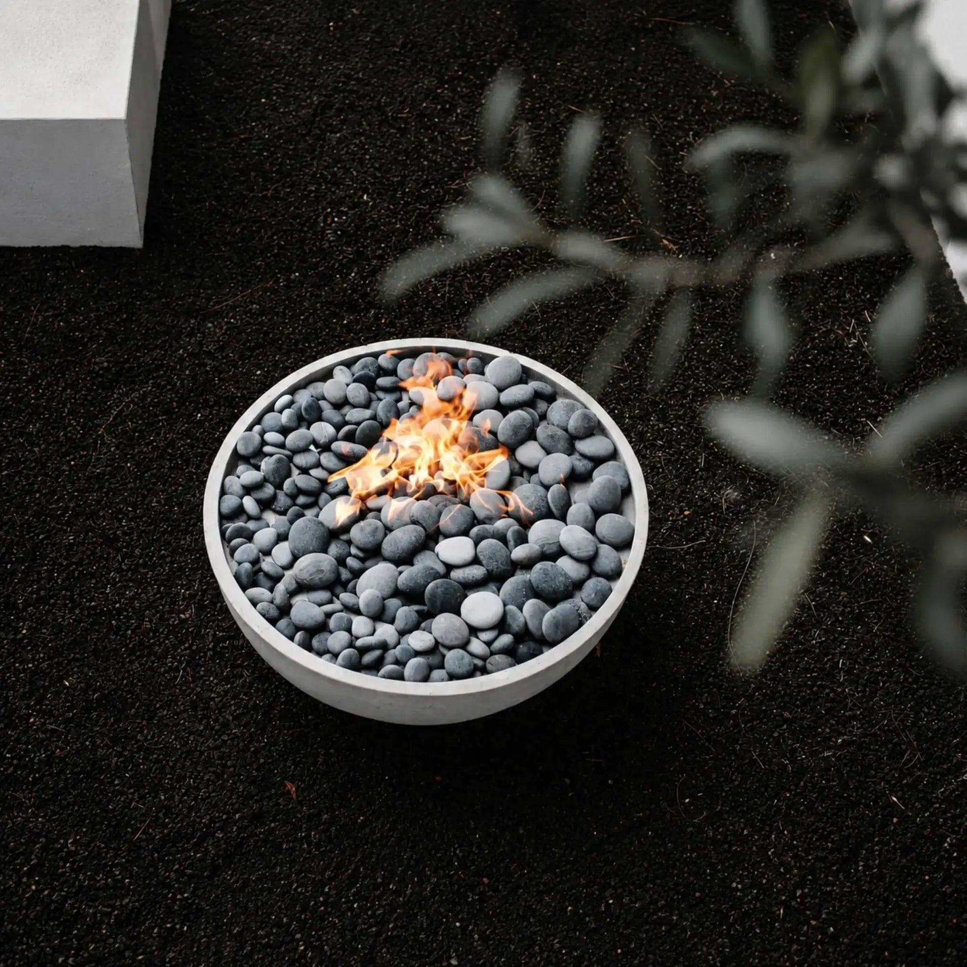HPC Fire Hearth Products Controls Dekko GFRC Concrete Serenade 32 Fire Pit displayed in a modern landscape setting with smooth gray fire stones and a natural gas flame burning at the center. The round concrete fire bowl sits on dark decorative gravel, viewed from above, while soft-focus greenery frames the foreground. This lifestyle image highlights the minimalist design and contemporary outdoor ambiance of the Serenade 32 Fire Pit.