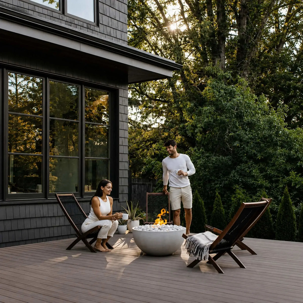 HPC Fire Hearth Products Controls Dekko GFRC Concrete Serenade 32 Fire Pit featured on a spacious backyard deck outside a modern home with dark siding and large black-framed windows. A couple relaxes around the round GFRC concrete fire bowl filled with white decorative stones and a warm flame. Surrounded by contemporary lounge chairs and mature trees, this lifestyle image showcases the Serenade 32 Fire Pit as an inviting focal point for outdoor entertaining and relaxation.