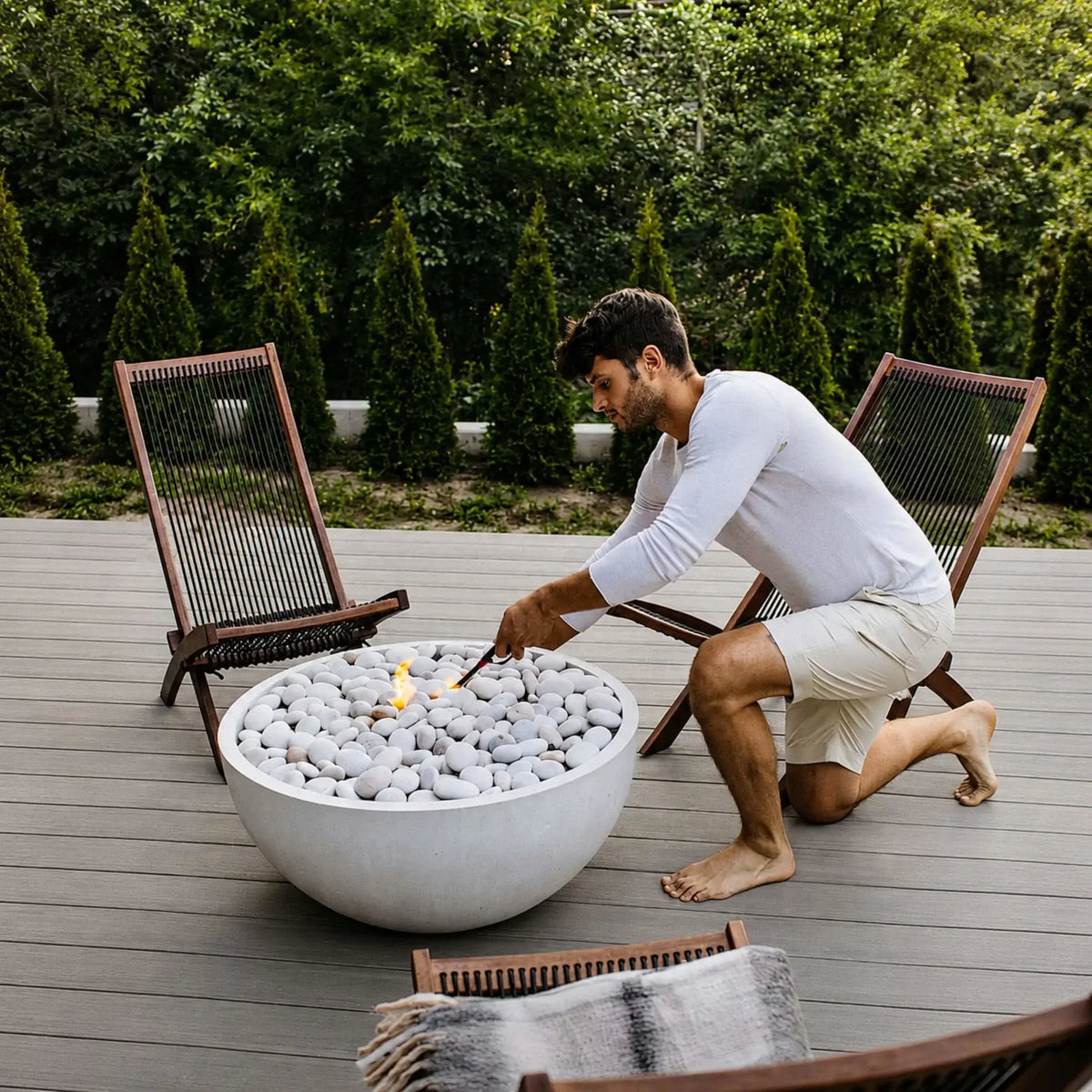 HPC Fire Hearth Products Controls Dekko GFRC Concrete Serenade 32 Fire Pit shown in use on a modern composite deck as a homeowner lights the burner. The round concrete fire bowl is filled with smooth white stones and positioned between contemporary outdoor lounge chairs. This lifestyle image highlights the hands-on operation, ease of ignition, and elegant design of the Serenade 32 Fire Pit in a residential outdoor living space.