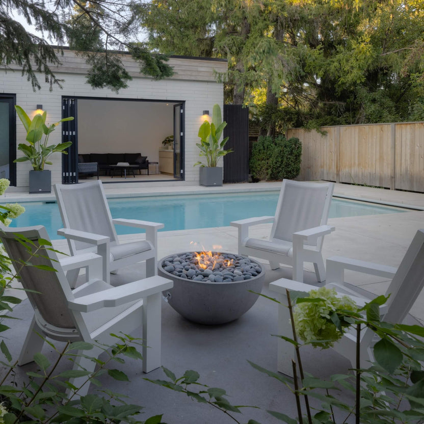 HPC Fire Hearth Products Controls Dekko GFRC Concrete Serenade 32 Fire Pit installed beside a backyard swimming pool, creating a cozy seating area with four white Adirondack chairs. The round fire bowl features gray decorative stones and a soft flame, surrounded by landscaped greenery and a modern pool house with folding glass doors. This lifestyle image demonstrates the Serenade 32 Fire Pit as a stylish centerpiece for poolside gatherings.