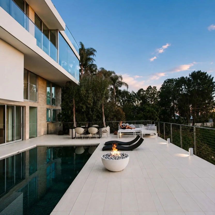 HPC Fire Hearth Products Controls Dekko GFRC Concrete Serenade 32 Fire Pit positioned beside a luxury swimming pool at a contemporary hillside residence. The round concrete fire bowl features decorative stone media and a bright flame, creating a focal point on the expansive patio. Surrounded by lounge seating, glass railings, and modern architecture, this lifestyle image showcases the Serenade 32 Fire Pit in an upscale outdoor living environment.