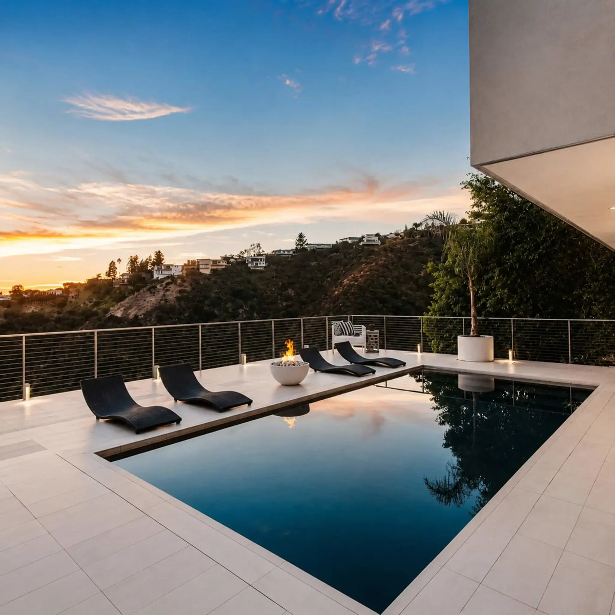 HPC Fire Hearth Products Controls Dekko GFRC Concrete Serenade 32 Fire Pit featured on a luxury rooftop-style pool terrace overlooking rolling hills at sunset. The round concrete fire bowl sits between modern chaise lounges, producing a warm flame that complements the tranquil infinity-style pool. This lifestyle image showcases the Serenade 32 Fire Pit as a stylish centerpiece for contemporary outdoor living spaces.