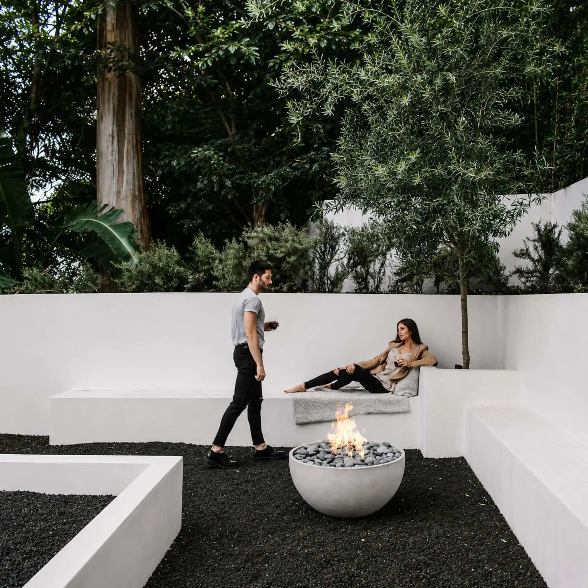 HPC Fire Hearth Products Controls Dekko GFRC Concrete Serenade 32 Fire Pit installed in a modern courtyard with clean white architectural walls and dark gravel landscaping. A couple relaxes near the round concrete fire bowl while flames rise above smooth gray stone media. This lifestyle image highlights the Serenade 32 Fire Pit as a sophisticated focal point for minimalist outdoor lounge and entertaining spaces.