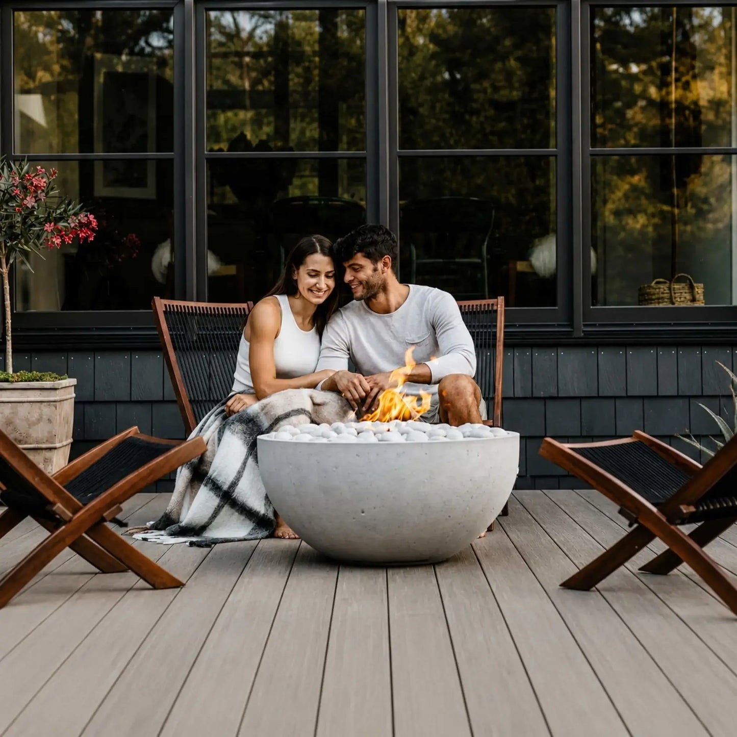 HPC Fire Hearth Products Controls Dekko GFRC Concrete Serenade 32 Fire Pit centered on a contemporary wood deck where a couple enjoys an intimate evening beside the fire. The round GFRC concrete bowl is filled with ivory stones and features a warm flame, surrounded by modern outdoor lounge chairs and large black-framed windows. This lifestyle image emphasizes comfort, relaxation, and stylish backyard entertaining.