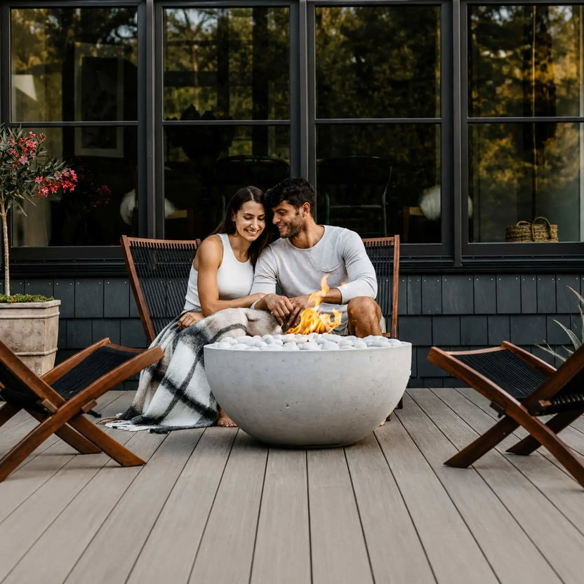 HPC Fire Hearth Products Controls Dekko GFRC Concrete Serenade 32 Fire Pit centered on a contemporary wood deck where a couple enjoys an intimate evening beside the fire. The round GFRC concrete bowl is filled with ivory stones and features a warm flame, surrounded by modern outdoor lounge chairs and large black-framed windows. This lifestyle image emphasizes comfort, relaxation, and stylish backyard entertaining.