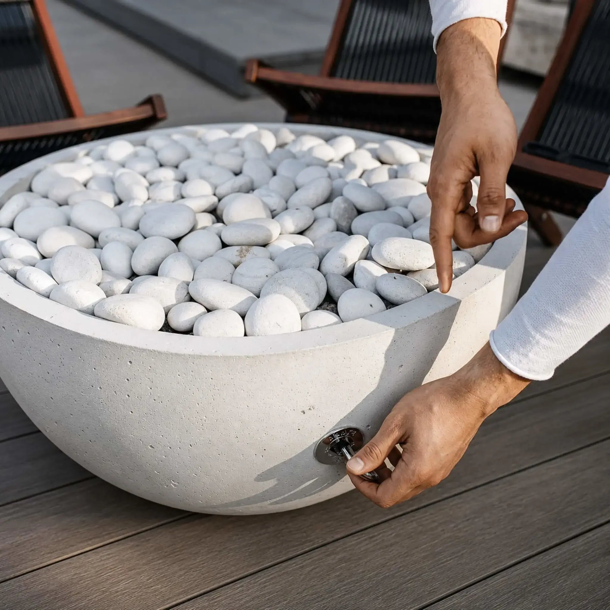 HPC Fire Hearth Products Controls Dekko GFRC Concrete Serenade 32 Fire Pit close-up showing the manual key valve operation on the side of the concrete bowl. A person’s hands demonstrate how to control the gas valve while the fire pit is filled with smooth ivory stones. Positioned on a modern deck with lounge seating in the background, this image highlights the functionality and user-friendly operation of the Serenade 32 Fire Pit.