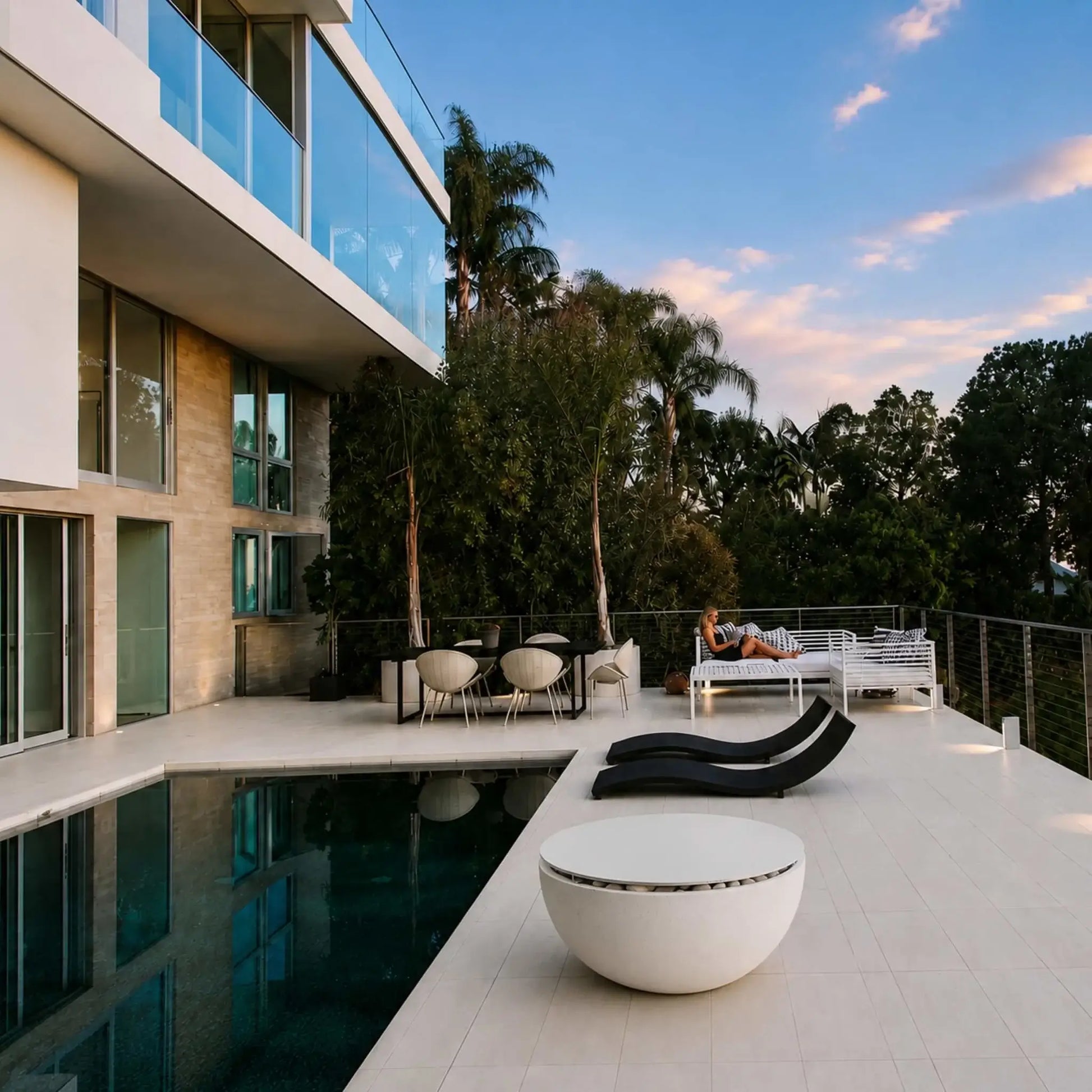 HPC Fire Hearth Products Controls Dekko GFRC Concrete Serenade 32 Fire Pit Table Top Lid featured beside a luxury modern swimming pool. The covered fire pit functions as a stylish outdoor table on a spacious patio surrounded by contemporary architecture, glass balcony railings, lounge seating, and tropical landscaping. This lifestyle image demonstrates how the Serenade 32 tabletop lid blends seamlessly into upscale outdoor living and entertainment spaces.
