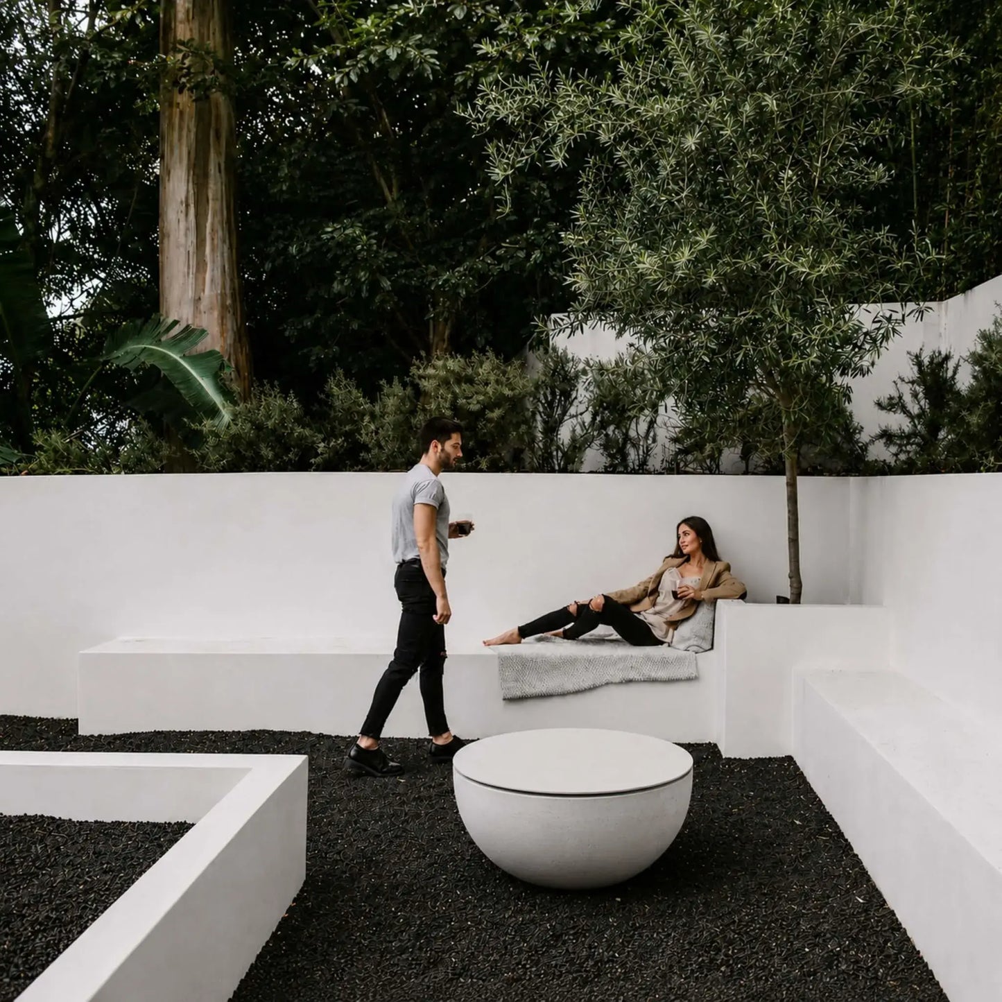 HPC Fire Hearth Products Controls Dekko GFRC Concrete Serenade 32 Fire Pit Table Top Lid displayed in a contemporary courtyard landscape. The covered round fire pit serves as a minimalist outdoor table surrounded by modern white concrete seating, decorative black stone ground cover, mature trees, and architectural garden walls. This lifestyle scene showcases the Serenade 32 tabletop lid as a sophisticated addition to modern outdoor entertaining spaces.