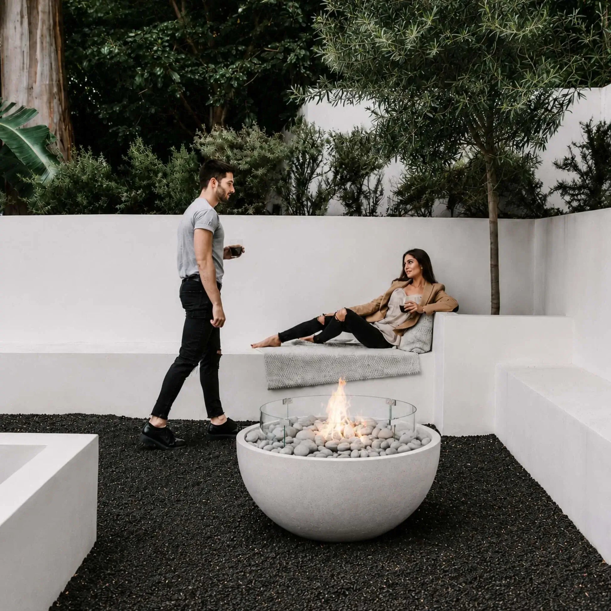 HPC Fire Hearth Products Controls Dekko GFRC Concrete Serenade 32 Fire Pit Tempered Glass Wind Guard Lifestyle 1 installation showing a modern outdoor lounge with a lit round fire bowl filled with smooth stones and protected by a clear glass wind guard. A man and woman relax beside the fire feature in a contemporary courtyard surrounded by white architectural seating, mature greenery, and elegant landscape design.