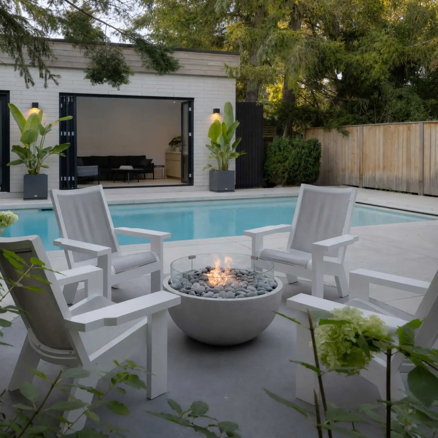 HPC Fire Hearth Products Controls Dekko GFRC Concrete Serenade 32 Fire Pit Tempered Glass Wind Guard Lifestyle 2 scene featuring a lit concrete fire bowl surrounded by white Adirondack-style chairs beside a modern swimming pool. The fire pit is filled with smooth gray stones and enclosed by a clear tempered glass wind guard, creating a welcoming outdoor gathering space with contemporary architecture, landscaping, and resort-style ambiance.
