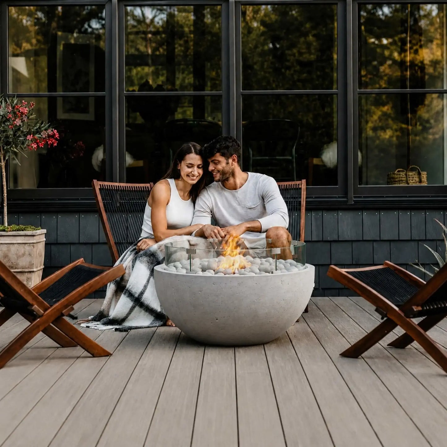 HPC Fire Hearth Products Controls Dekko GFRC Concrete Serenade 32 Fire Pit Tempered Glass Wind Guard Lifestyle 3 scene featuring a couple relaxing together on a modern outdoor deck beside a lit Serenade 32 fire pit. The round GFRC concrete fire bowl is filled with smooth decorative fire stones and protected by a clear tempered glass wind guard. Surrounded by contemporary wood lounge chairs and set against a dark modern home exterior with large windows, the fire feature creates a warm atmosphere.