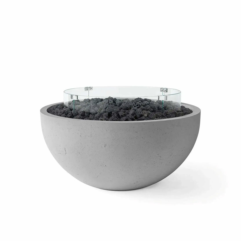 HPC Fire Hearth Products Controls Dekko GFRC Concrete Serenade 40 Fire Pit in Charcoal finish with black lava rock media and a clear tempered glass wind guard. This modern round GFRC concrete fire bowl features a rich charcoal-gray exterior, rugged volcanic rock fill, and a circular glass windscreen that enhances flame performance while maintaining a clean, contemporary outdoor aesthetic.