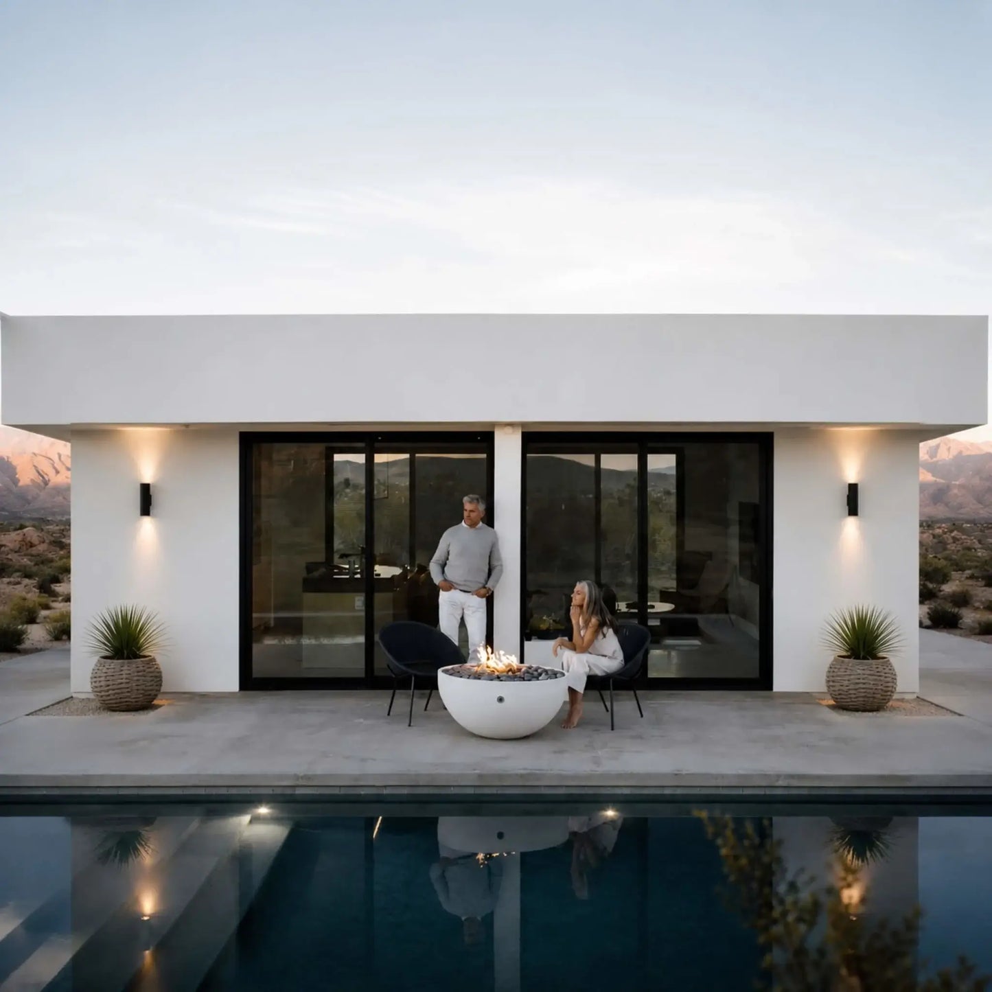 HPC Fire Hearth Products Controls Dekko GFRC Concrete Serenade 40 Fire Pit Lifestyle image showcasing a modern desert home with a white round Serenade 40 fire pit centered beside a reflective swimming pool. A couple relaxes around the glowing fire feature on a minimalist patio, highlighting the fire pit’s contemporary GFRC concrete construction, clean lines, and ability to create a warm outdoor gathering space in a luxury residential setting.