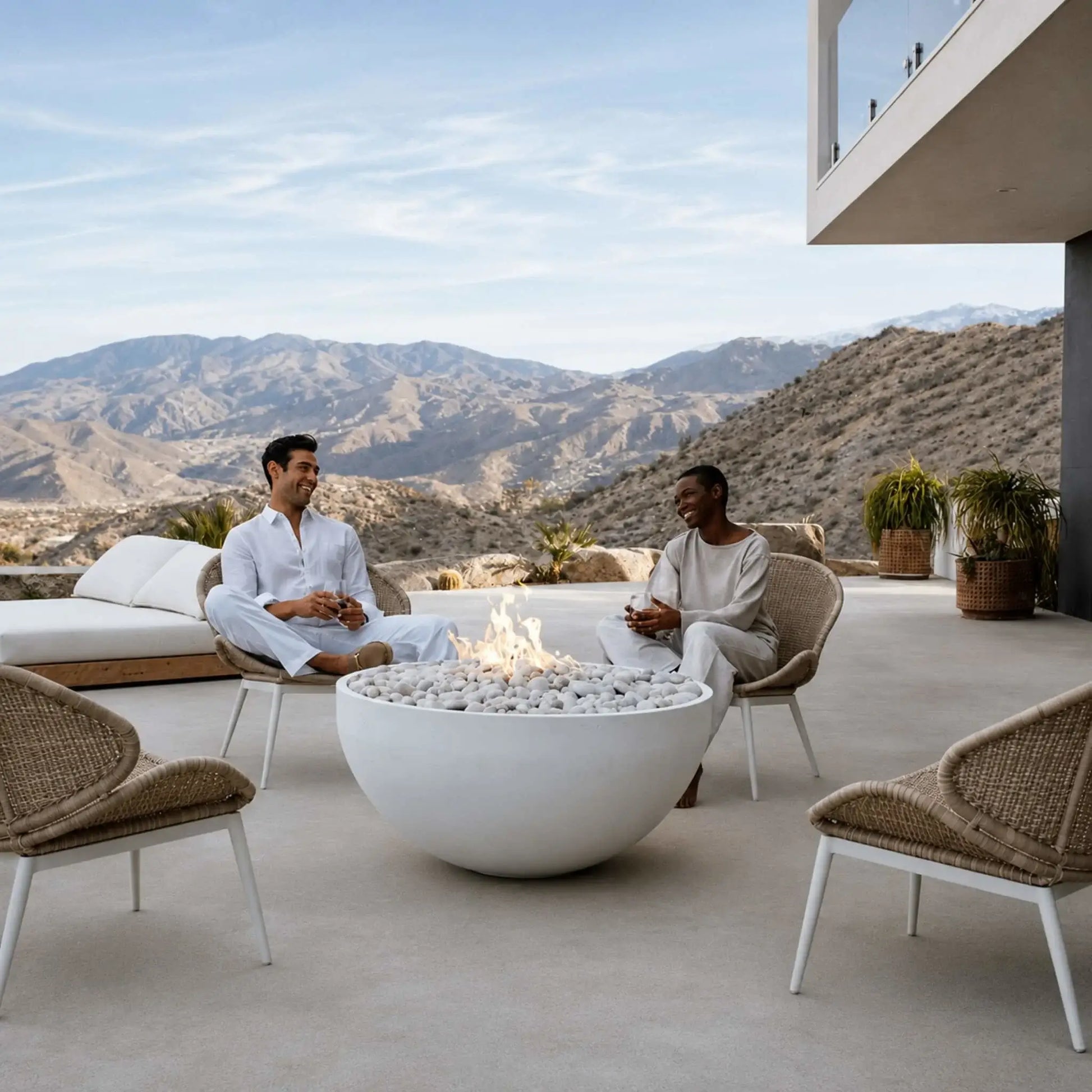 HPC Fire Hearth Products Controls Dekko GFRC Concrete Serenade 40 Fire Pit Lifestyle scene with two people relaxing around a white GFRC concrete fire bowl filled with decorative stone media. Set on a modern outdoor terrace overlooking desert mountains, the Serenade 40 fire pit creates a warm gathering space surrounded by woven patio chairs, luxury architecture, and expansive scenic views.