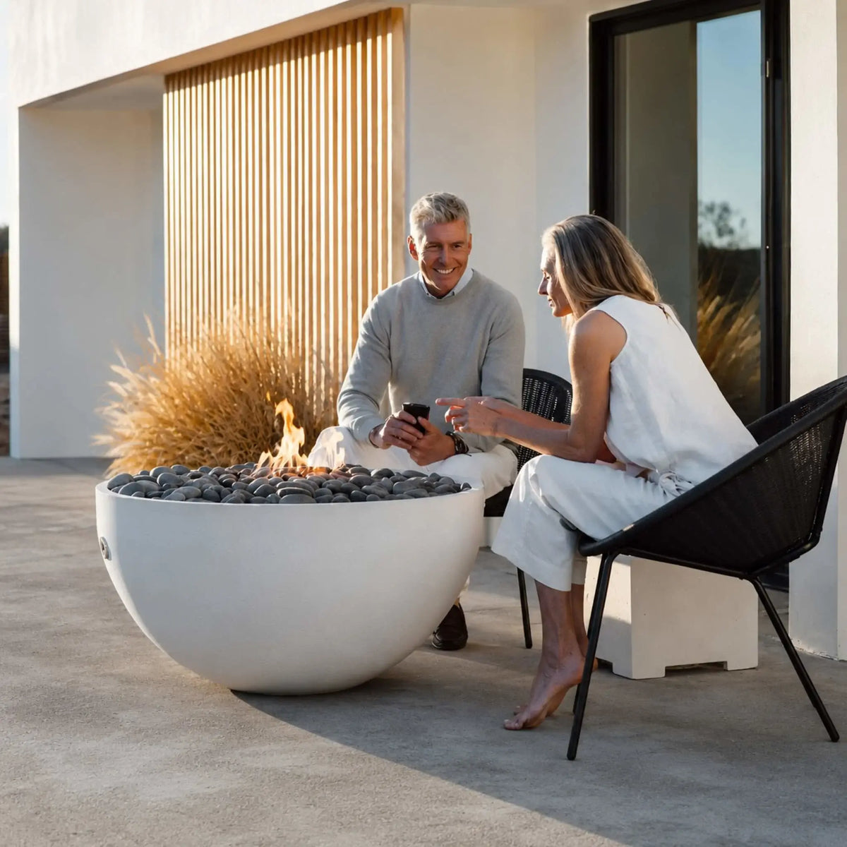HPC Fire Hearth Products Controls Dekko GFRC Concrete Serenade 40 Fire Pit Lifestyle image showing a modern white fire bowl filled with dark decorative stones and dancing flames beside a contemporary home. A couple sits comfortably in black outdoor lounge chairs enjoying the warmth of the Serenade 40 fire pit, highlighting its elegant design for luxury patios and outdoor living areas.