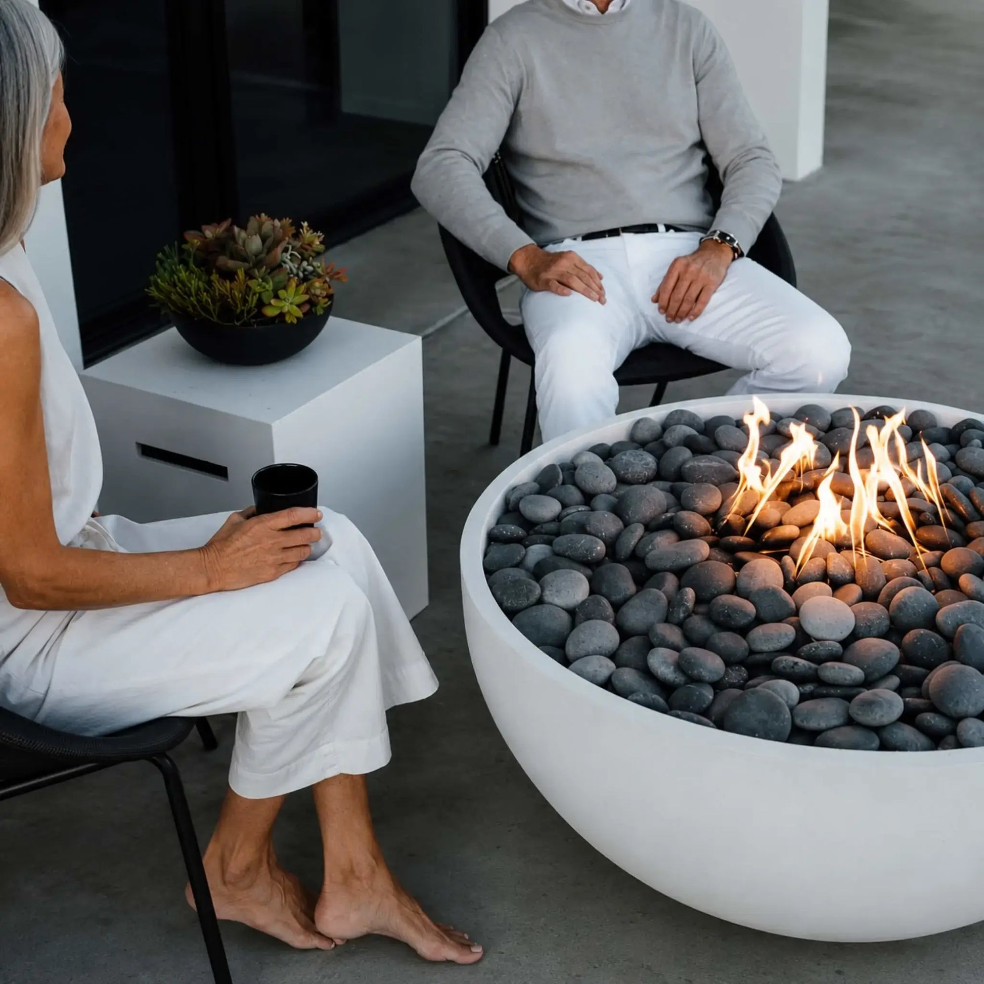 HPC Fire Hearth Products Controls Dekko GFRC Concrete Serenade 40 Fire Pit Lifestyle close-up featuring a round white concrete fire pit filled with smooth gray river stones and a bright gas flame. Seated guests enjoy drinks around the Serenade 40 fire pit on a modern patio, emphasizing the decorative fire media, premium craftsmanship, and inviting outdoor atmosphere.