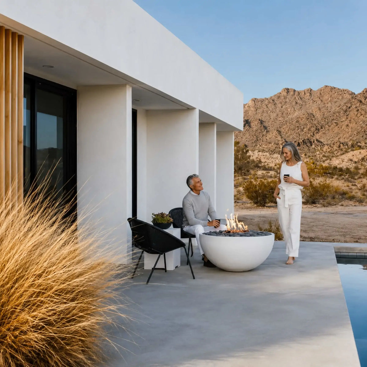 HPC Fire Hearth Products Controls Dekko GFRC Concrete Serenade 40 Fire Pit Lifestyle image showing a contemporary desert residence with a white round fire pit burning beside a pool. A couple enjoys the outdoor space surrounded by desert landscaping, mountain views, and modern architecture, highlighting the Serenade 40 GFRC concrete fire pit as a stylish backyard focal point.