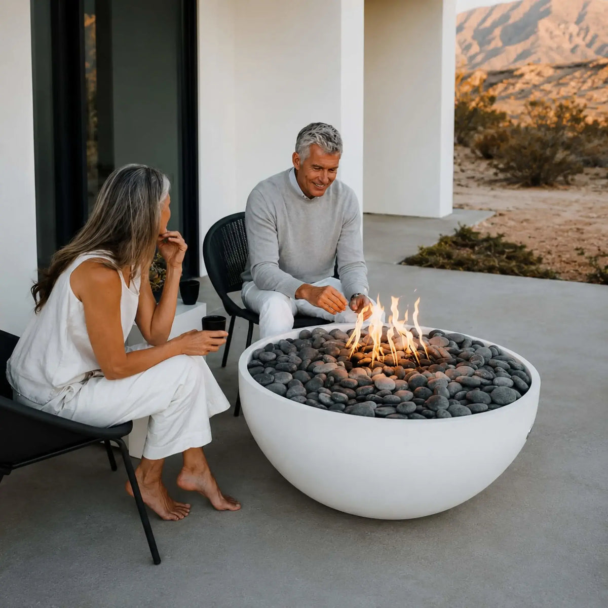 HPC Fire Hearth Products Controls Dekko GFRC Concrete Serenade 40 Fire Pit Lifestyle scene featuring a couple gathered around a white concrete fire bowl filled with smooth river stones. The Serenade 40 fire pit produces warm flames on a contemporary patio overlooking a desert landscape, showcasing its clean lines, durable GFRC construction, and upscale outdoor living appeal.