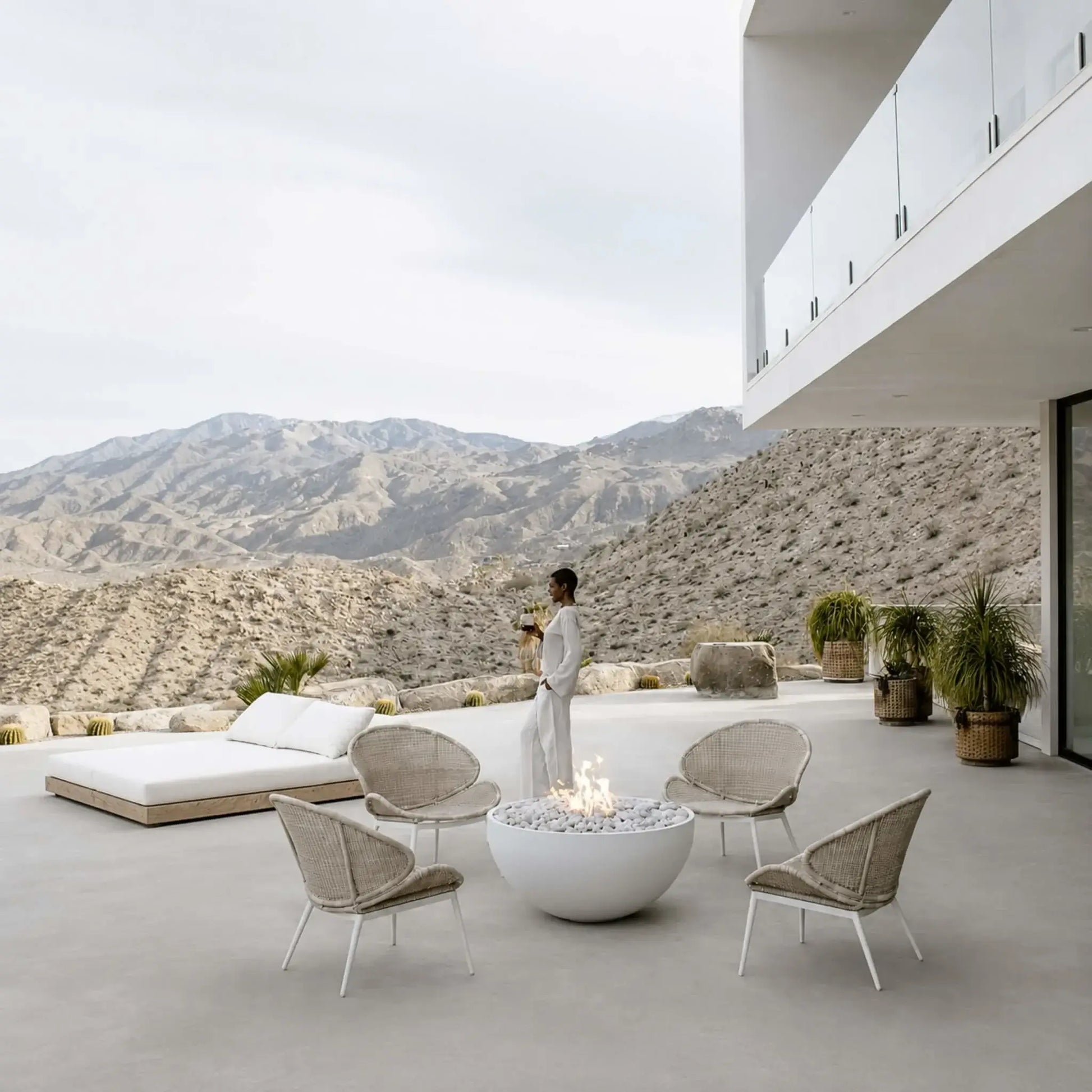 HPC Fire Hearth Products Controls Dekko GFRC Concrete Serenade 40 Fire Pit Lifestyle image featuring a luxury outdoor terrace with four woven lounge chairs arranged around a white concrete fire bowl. The Serenade 40 fire pit glows with flames over decorative stone media against a dramatic mountain backdrop, creating a sophisticated outdoor gathering space for entertaining and relaxation.