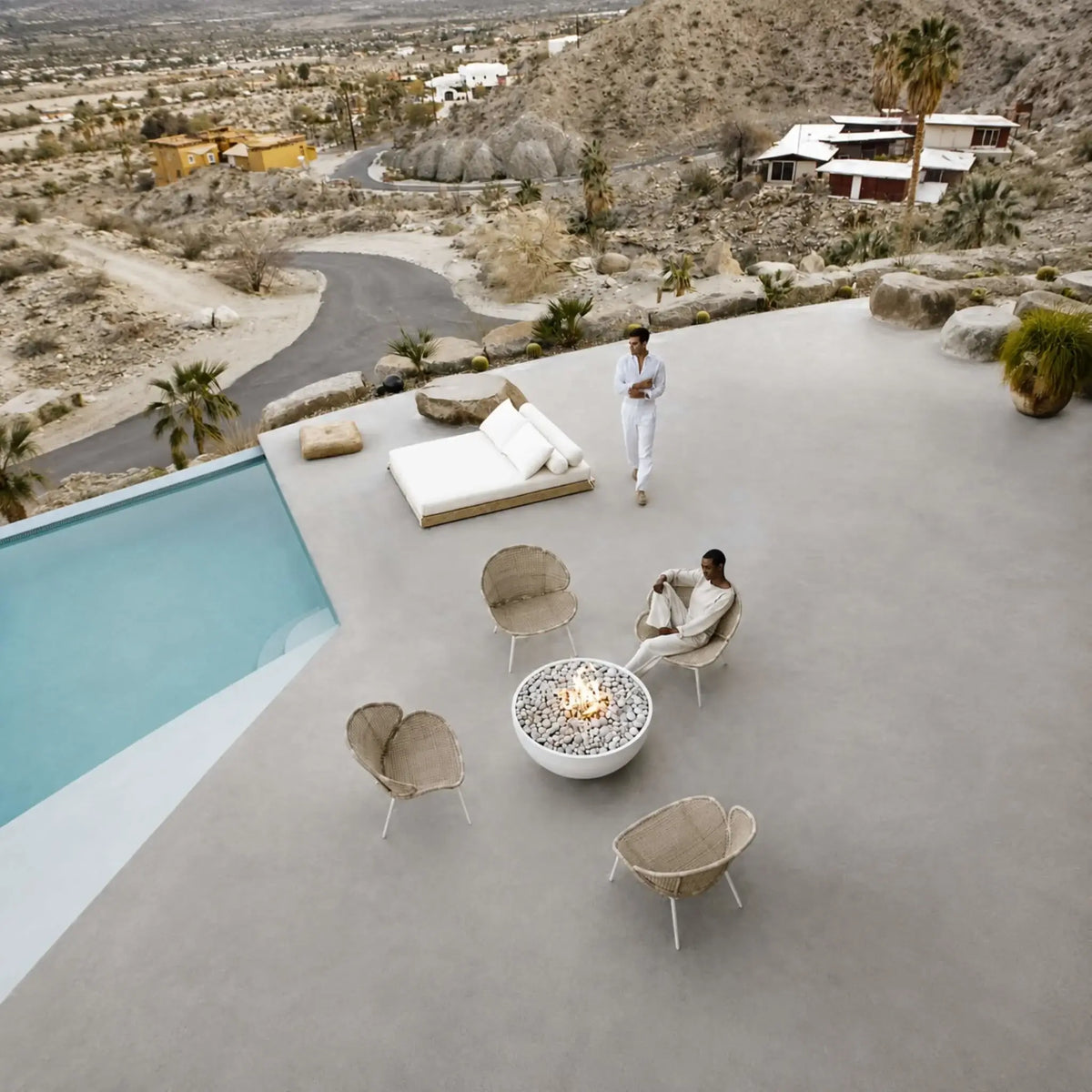 HPC Fire Hearth Products Controls Dekko GFRC Concrete Serenade 40 Fire Pit Lifestyle aerial view showing a modern hillside patio with a white round fire pit, woven lounge seating, infinity-edge pool, and expansive desert landscape. The Serenade 40 GFRC concrete fire pit serves as the focal point of the outdoor living area, blending contemporary design with luxury resort-style surroundings.