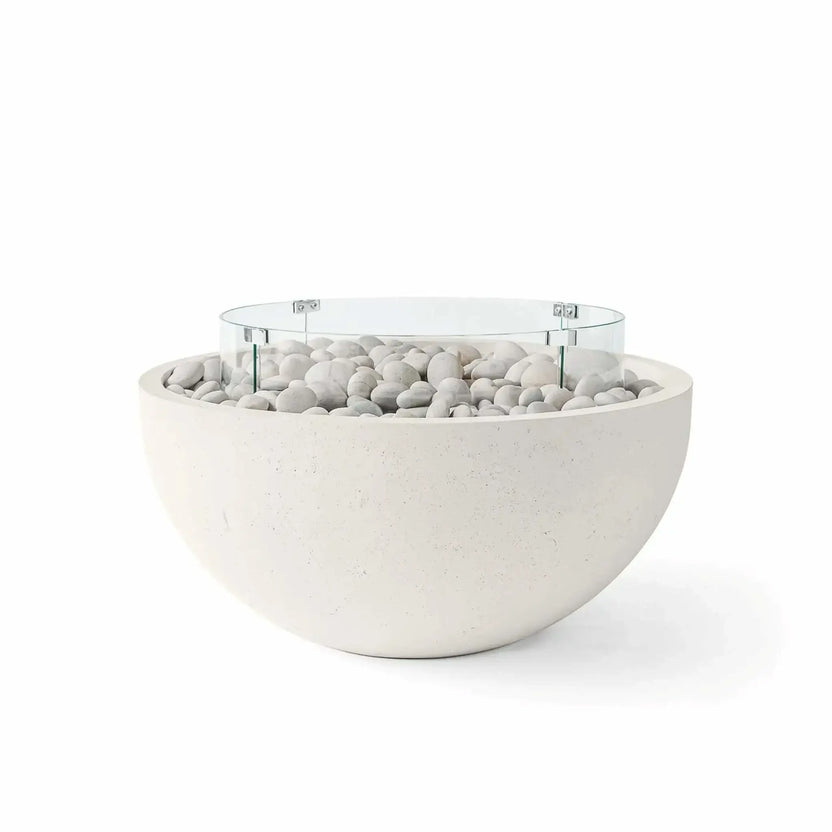HPC Fire Hearth Products Controls Dekko GFRC Concrete Serenade 40 Fire Pit in Limestone finish with ivory stone fire media and a tempered glass wind guard. The light limestone-colored GFRC concrete bowl features decorative white river stones, a circular tempered glass flame barrier, and a contemporary rounded silhouette, creating a bright, elegant outdoor fire pit centerpiece for luxury patios and modern landscape settings.