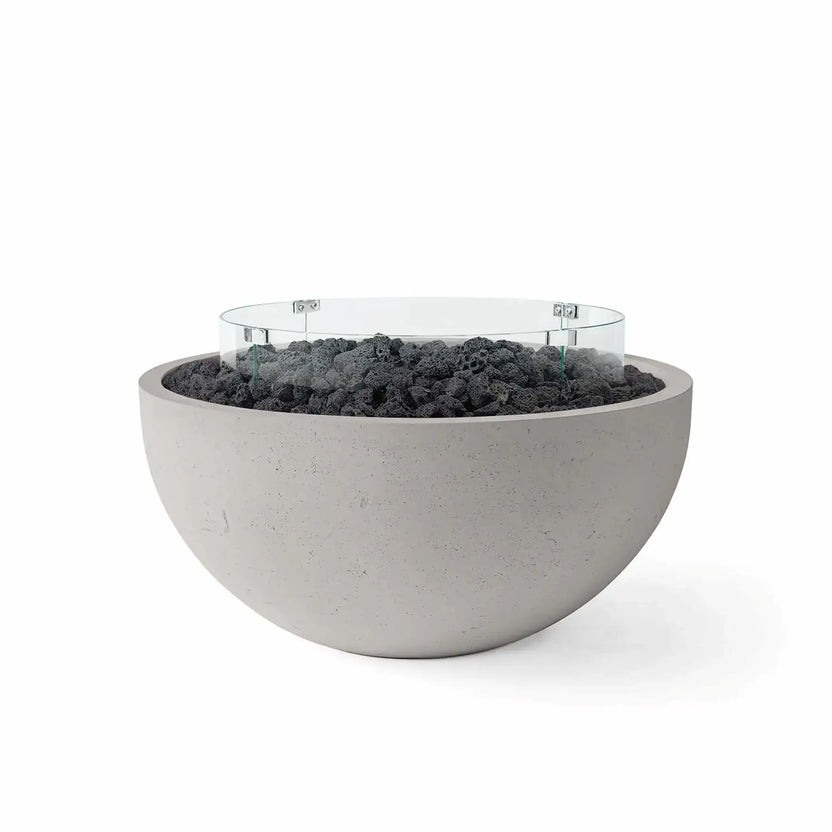 HPC Fire Hearth Products Controls Dekko GFRC Concrete Serenade 40 Fire Pit in Sandstone finish featuring black lava rock fire media and a clear tempered glass wind guard. The rounded concrete fire bowl pairs a warm neutral exterior with dark volcanic rock for dramatic contrast, while the circular glass windscreen enhances flame control and visual appeal in modern outdoor living spaces.