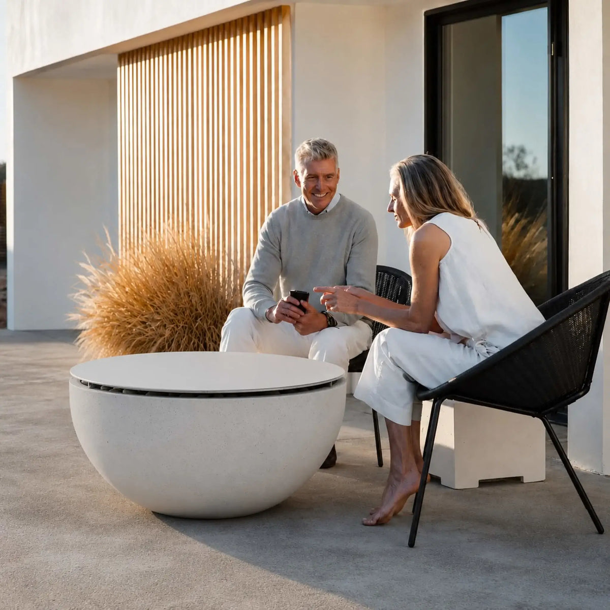 HPC Fire Hearth Products Controls Dekko GFRC Concrete Serenade 40 Fire Pit Table Top Lid showcased in an upscale residential outdoor living area. A couple relaxes beside the covered GFRC concrete fire pit, which serves as a stylish coffee table with its round tabletop lid in place. The modern patio design, neutral furnishings, and warm architectural elements highlight the fire pit’s versatility and refined appearance.