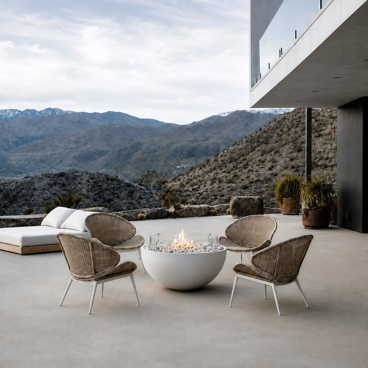 HPC Fire Hearth Products Controls Dekko GFRC Concrete Serenade 40 Fire Pit Tempered Glass Wind Guard Lifestyle 3 image showcasing a luxury outdoor patio with panoramic mountain views. Centered in a contemporary seating arrangement, the round white GFRC concrete Serenade 40 fire pit features ivory stone fire media, a tempered glass wind guard, and a glowing flame. Woven lounge chairs, a modern hillside residence, and expansive open-air architecture create an elegant outdoor living space.