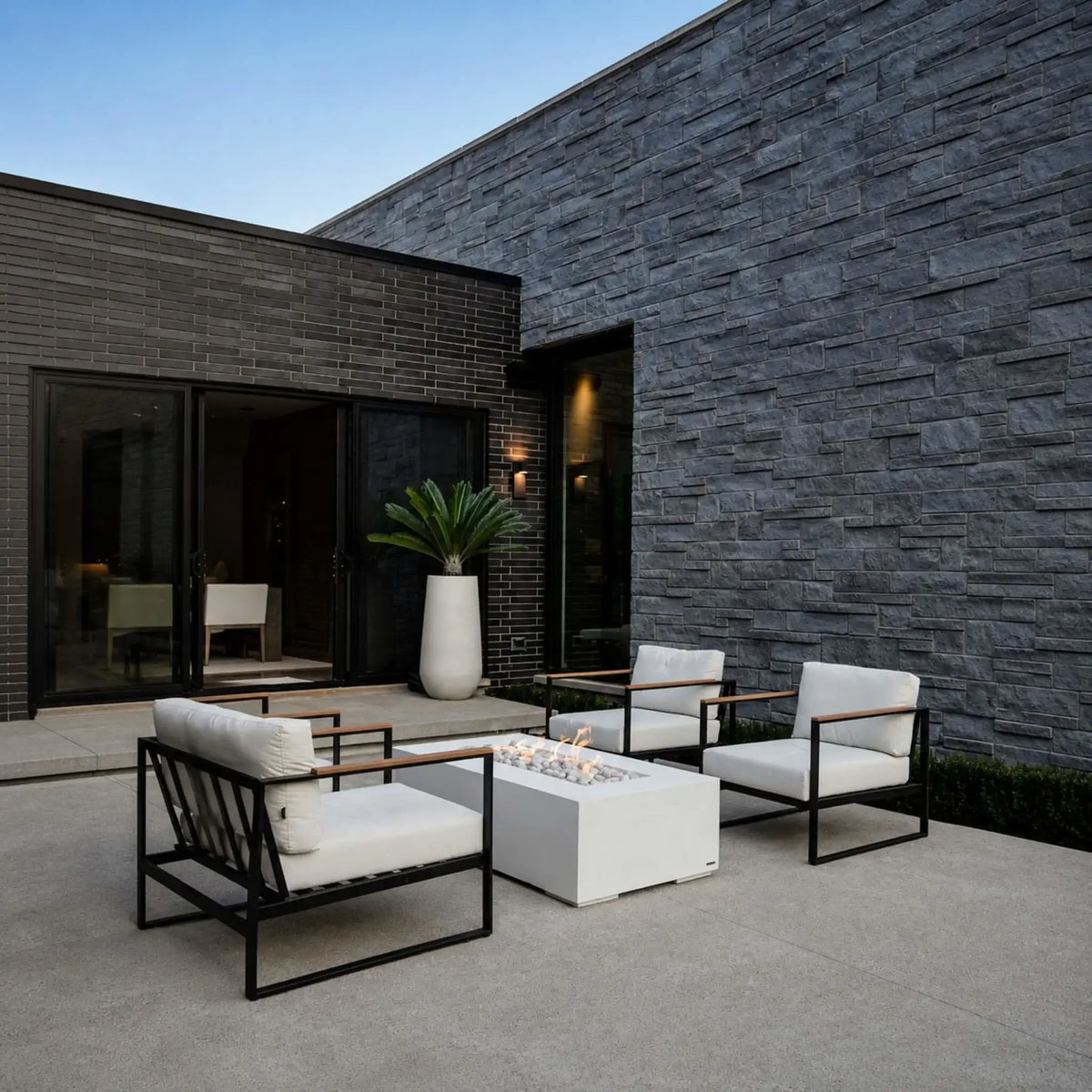 HPC Fire Hearth Products Controls Dekko GFRC Concrete Sonoma 56 x 30 Fire Pit showcased in a modern outdoor courtyard with contemporary lounge seating. The white rectangular Sonoma fire pit serves as the focal point between black-framed chairs with white cushions, surrounded by dark brick and stacked stone walls. Soft flames rise from decorative ivory stones, creating a sophisticated outdoor living environment ideal for entertaining and relaxation.