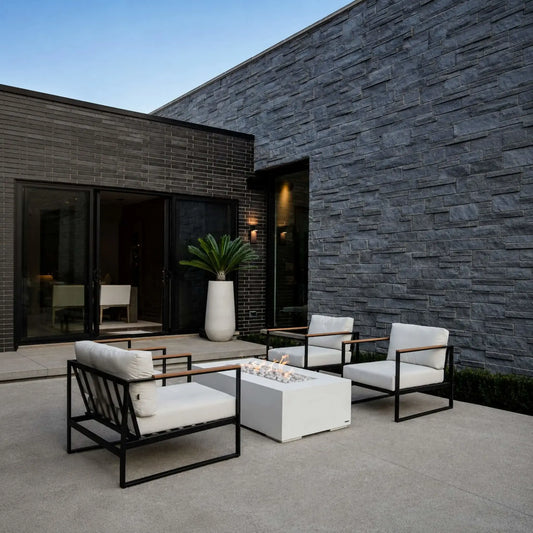 HPC Fire Hearth Products Controls Dekko GFRC Concrete Sonoma 56 x 30 Fire Pit showcased in a modern outdoor courtyard with contemporary lounge seating. The white rectangular Sonoma fire pit serves as the focal point between black-framed chairs with white cushions, surrounded by dark brick and stacked stone walls. Soft flames rise from decorative ivory stones, creating a sophisticated outdoor living environment ideal for entertaining and relaxation.