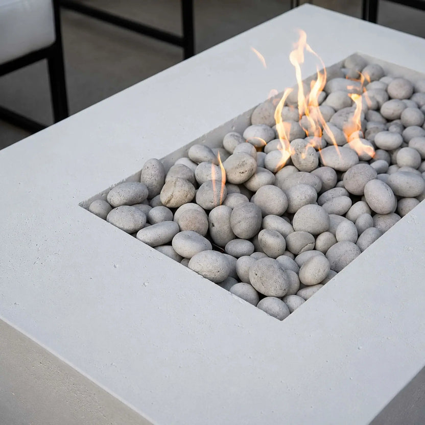Close-up lifestyle image of the HPC Fire Hearth Products Controls Dekko GFRC Concrete Sonoma 56 x 30 Fire Pit. The recessed burner area is filled with smooth ivory fire stones while natural gas flames dance across the surface. The white GFRC concrete tabletop highlights the clean lines and contemporary design of the Sonoma fire pit, emphasizing premium materials and outdoor luxury patio styling.