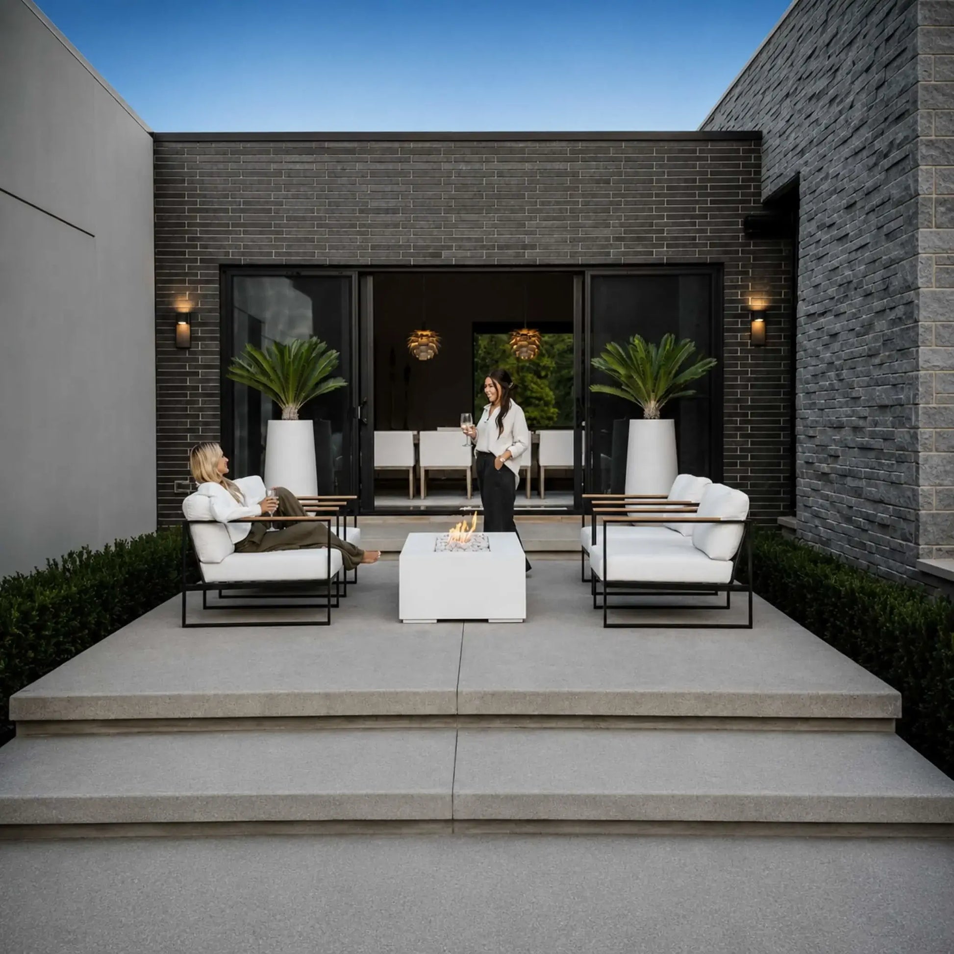 HPC Fire Hearth Products Controls Dekko GFRC Concrete Sonoma 56 x 30 Fire Pit featured in a luxury residential patio setting. Two people enjoy a contemporary outdoor seating area centered around the white rectangular Sonoma fire pit with decorative stone media and visible flames. The modern courtyard includes architectural stone walls, large glass doors, landscape lighting, and minimalist outdoor furnishings.