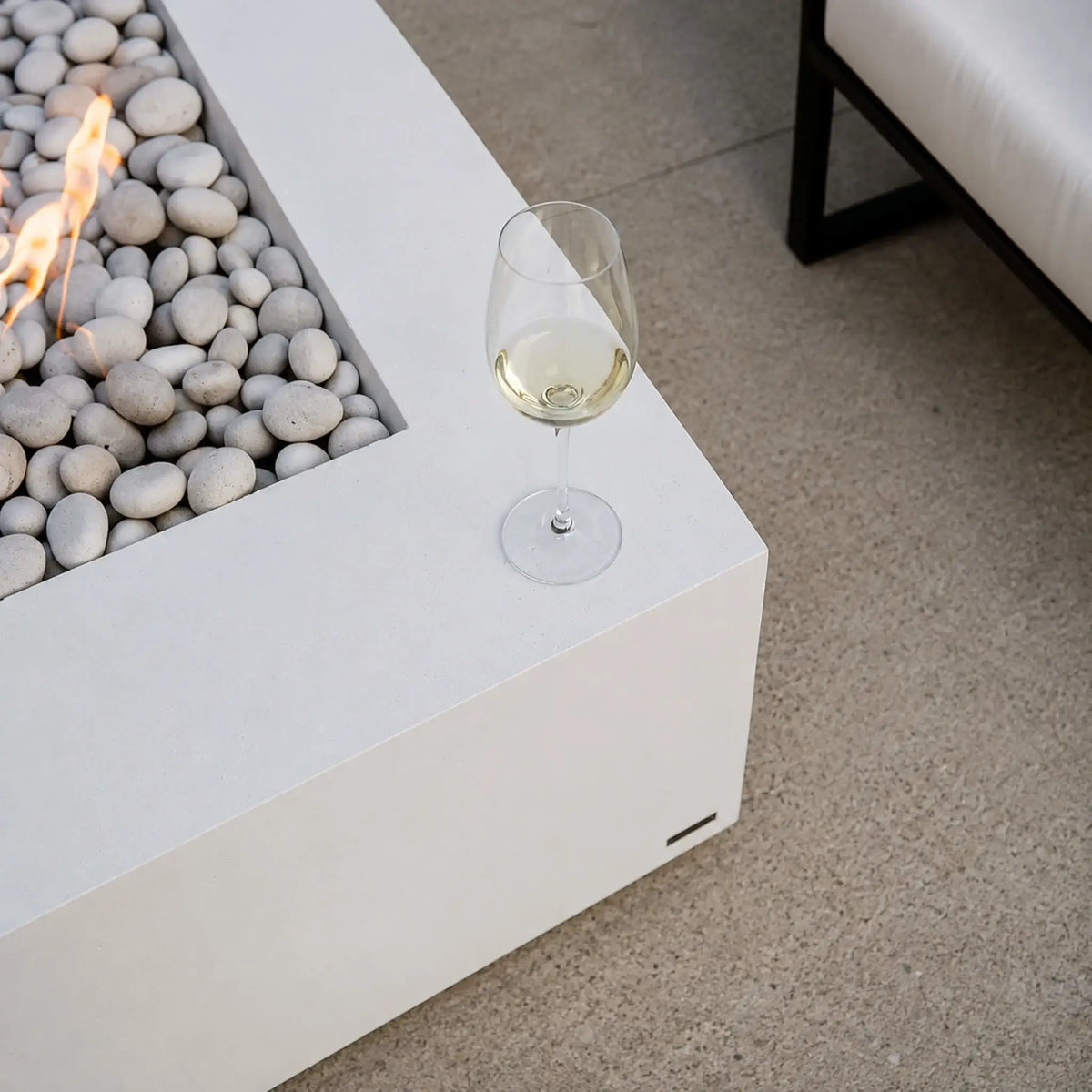 Detail view of the HPC Fire Hearth Products Controls Dekko GFRC Concrete Sonoma 56 x 30 Fire Pit showing the corner of the white concrete fire table with ivory stone media and active flames. A glass of white wine rests on the tabletop edge, highlighting the fire pit’s functional surface area and entertaining appeal. This close-up lifestyle image showcases the Sonoma fire pit’s clean design and upscale outdoor living aesthetic.