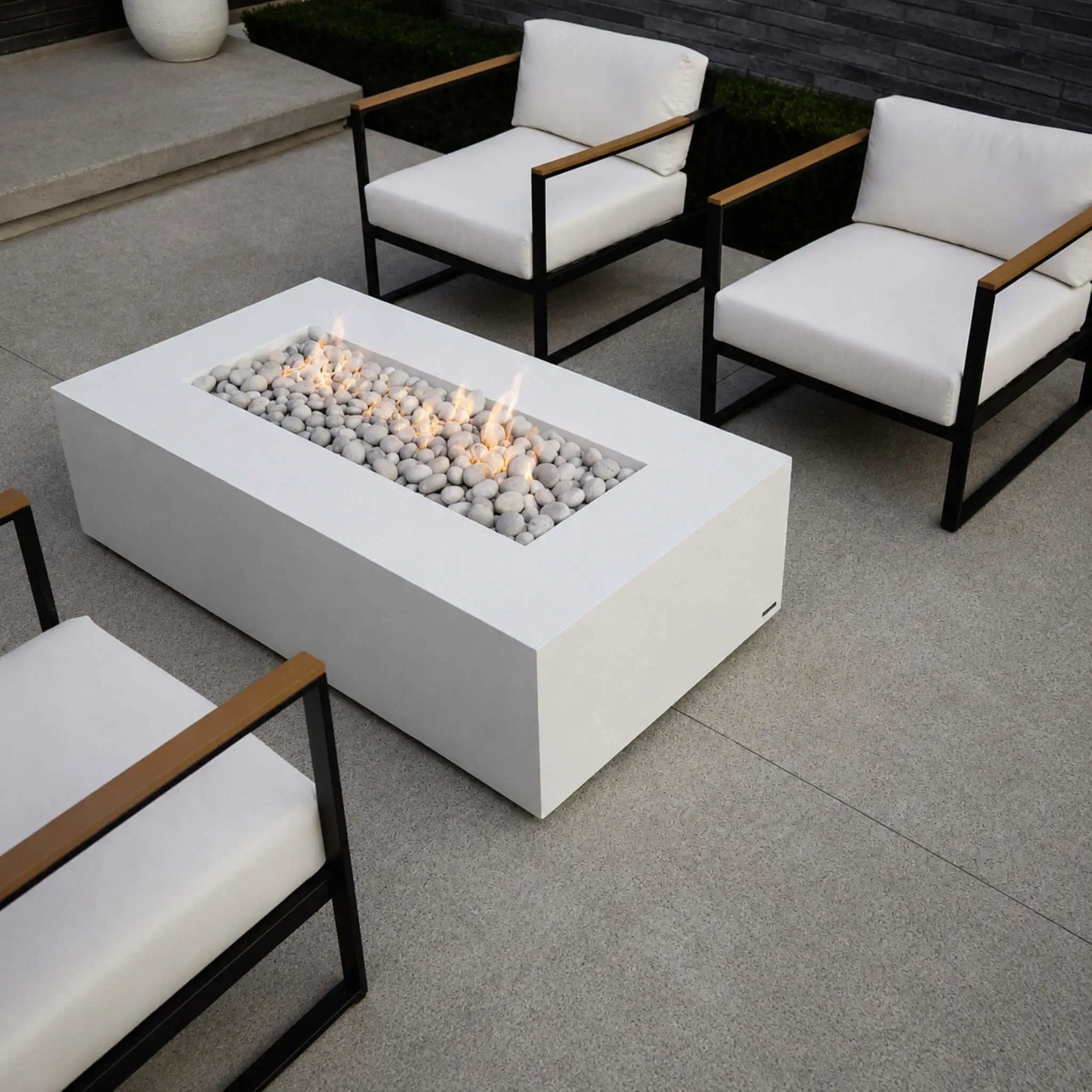 Elevated lifestyle view of the HPC Fire Hearth Products Controls Dekko GFRC Concrete Sonoma 56 x 30 Fire Pit positioned between modern outdoor lounge chairs. The white rectangular GFRC fire pit features a linear burner filled with ivory stones and glowing flames, creating a welcoming gathering space. This contemporary patio scene highlights the Sonoma fire pit as a stylish centerpiece for luxury outdoor entertaining areas.