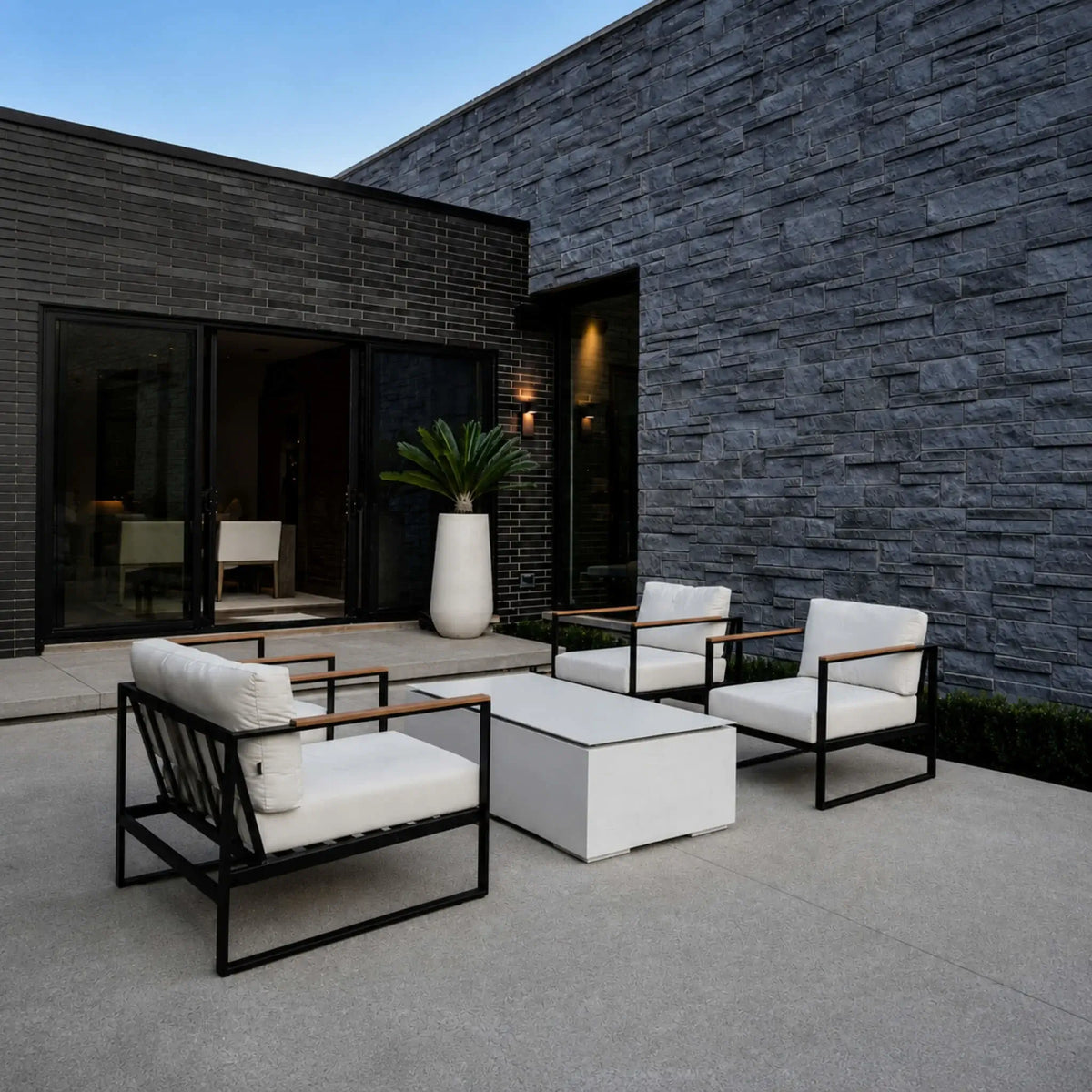 HPC Fire Hearth Products Controls Dekko GFRC Concrete Sonoma 56 x 30 Fire Pit Table Top Lid Lifestyle 1 image featuring the covered fire pit in a modern outdoor seating area. The rectangular tabletop lid transforms the fire pit into a stylish coffee table surrounded by black metal lounge chairs with white cushions. Set against contemporary stone and brick architecture, the scene highlights the versatility of the Sonoma fire pit when not in use as a fire feature.