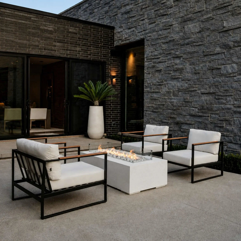 HPC Fire Hearth Products Controls Dekko GFRC Concrete Sonoma 56 x 30 Fire Pit Tempered Glass Wind Guard Lifestyle 1 image showing the modern rectangular fire pit installed in a luxury outdoor seating area. The white GFRC concrete fire pit sits between contemporary black metal lounge chairs with white cushions, featuring a linear flame display protected by a tempered glass wind guard. Set against dark brick and stone architecture, the scene highlights an upscale outdoor living and entertaining environment.