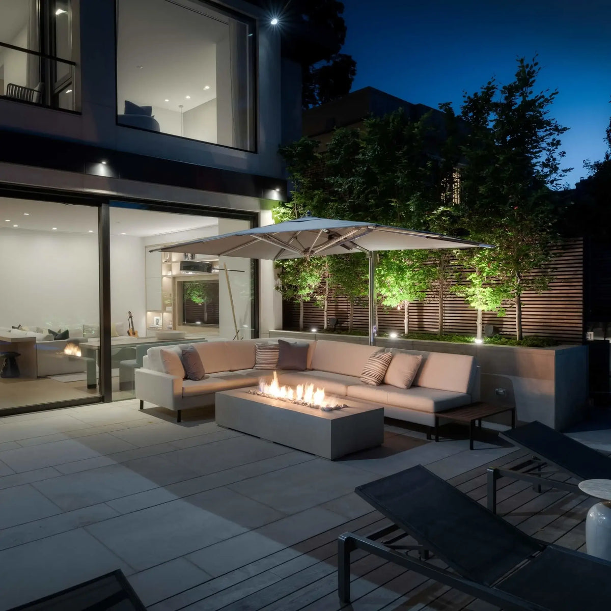 The Dekko GFRC Concrete Sonoma 72 x 33 Fire Pit serves as the centerpiece of a luxurious modern outdoor living space at night. Positioned beneath a large patio umbrella and surrounded by contemporary sectional seating, the rectangular fire pit features a vibrant linear flame that creates a warm gathering area. Floor-to-ceiling glass walls, architectural lighting, and landscaped greenery showcase the Sonoma fire pit’s sleek design and upscale residential appeal.