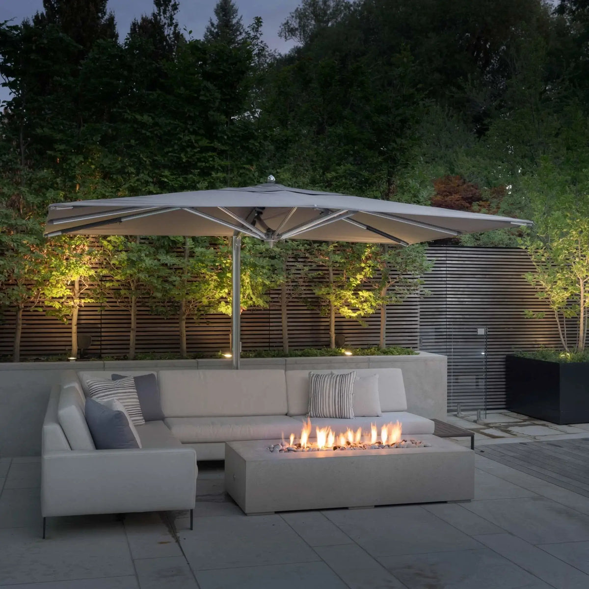 Close-up lifestyle image of the Dekko GFRC Concrete Sonoma 72 x 33 Fire Pit installed on a modern patio. The rectangular concrete fire feature produces a continuous linear flame above decorative fire media and is paired with a contemporary outdoor sectional beneath a large cantilever umbrella. Illuminated trees, privacy screening, and clean architectural lines highlight the fire pit’s sophisticated outdoor entertaining style.