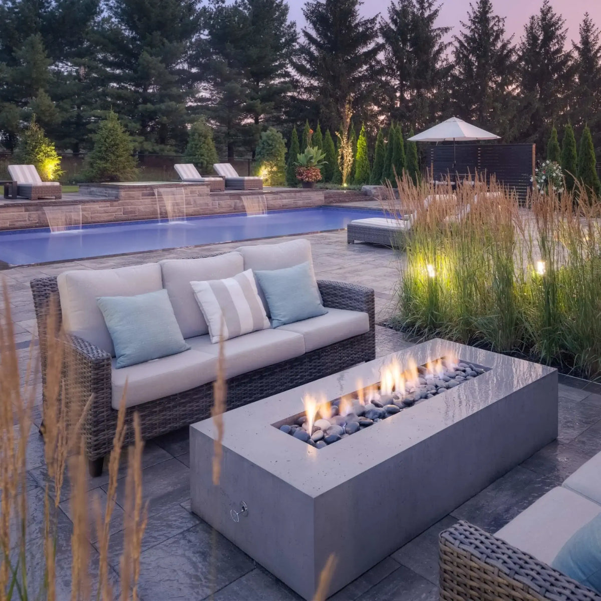 The Dekko Sonoma 72 x 33 GFRC Concrete Fire Pit is showcased beside an elegant resort-style swimming pool surrounded by lush landscaping and outdoor lounge seating. The rectangular fire pit features dancing flames over decorative stone media, creating a warm focal point among wicker furniture, ornamental grasses, and illuminated water features. This lifestyle setting highlights the fire pit’s versatility in upscale backyard retreats.