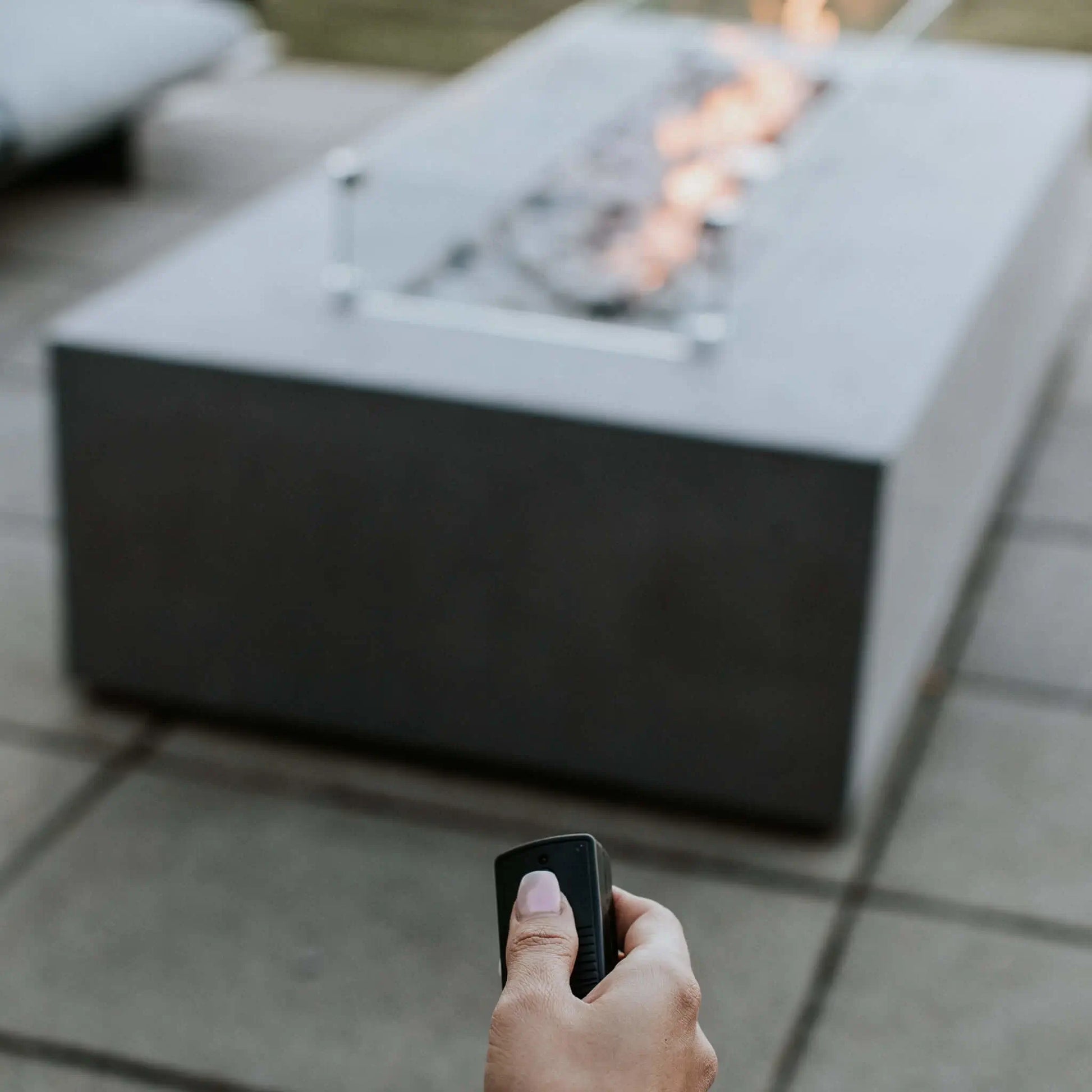 A hand operates a remote control aimed at the Dekko GFRC Concrete Sonoma 72 x 33 Fire Pit, demonstrating the convenience of electronic ignition and remote flame control. The rectangular fire pit burns steadily in the background with a linear flame protected by a glass wind guard. This close-up lifestyle image emphasizes the Sonoma fire pit’s user-friendly operation and modern outdoor functionality.