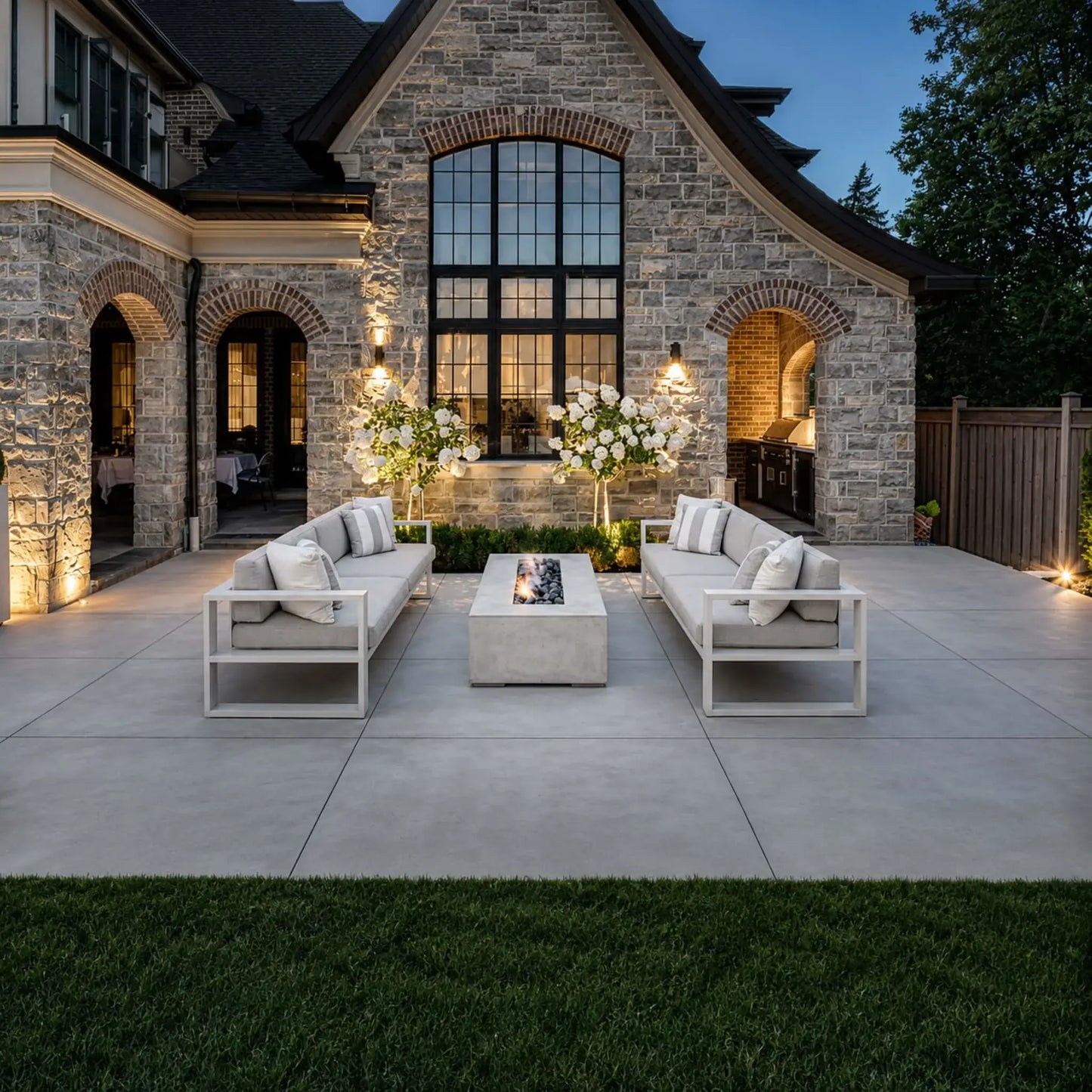 Evening courtyard scene showcasing the Dekko GFRC Concrete Sonoma 72 x 33 Fire Pit positioned between two outdoor sofas in front of an elegant stone estate home. The rectangular fire feature displays a glowing linear flame over dark fire media, creating a warm gathering space. Landscape lighting, architectural stonework, and luxury outdoor furnishings enhance the sophisticated ambiance.