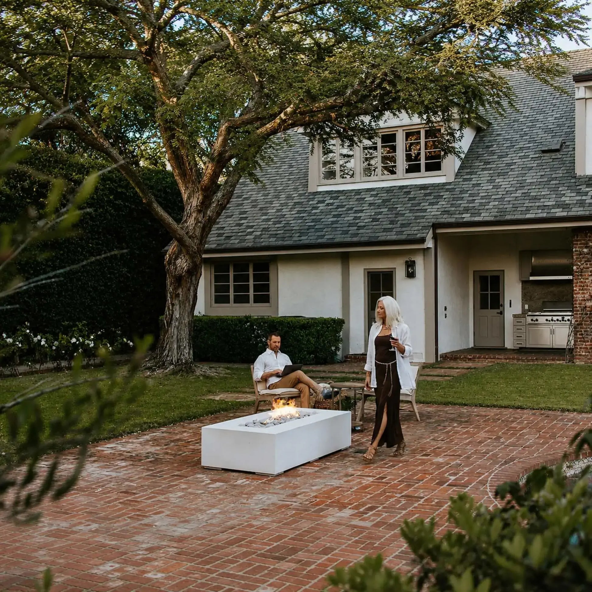 Dekko Sonoma 72 x 33 GFRC Concrete Fire Pit featured in a luxury backyard with a brick patio, mature shade tree, and swimming pool. A woman walks near the active fire pit while another person relaxes in nearby seating. The rectangular white concrete fire feature serves as a stylish centerpiece, adding warmth and modern design to the outdoor living environment.